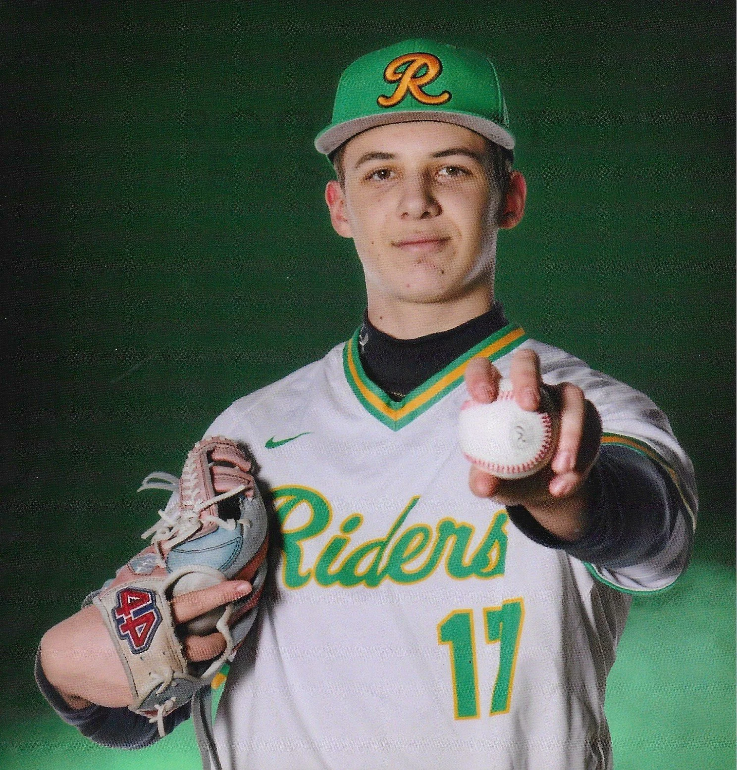 Left-handed pitcher male baseball player in a green hat and white jersey with green and yellow trim, pointing towards the camera, holding a baseball and glove, against a green background.