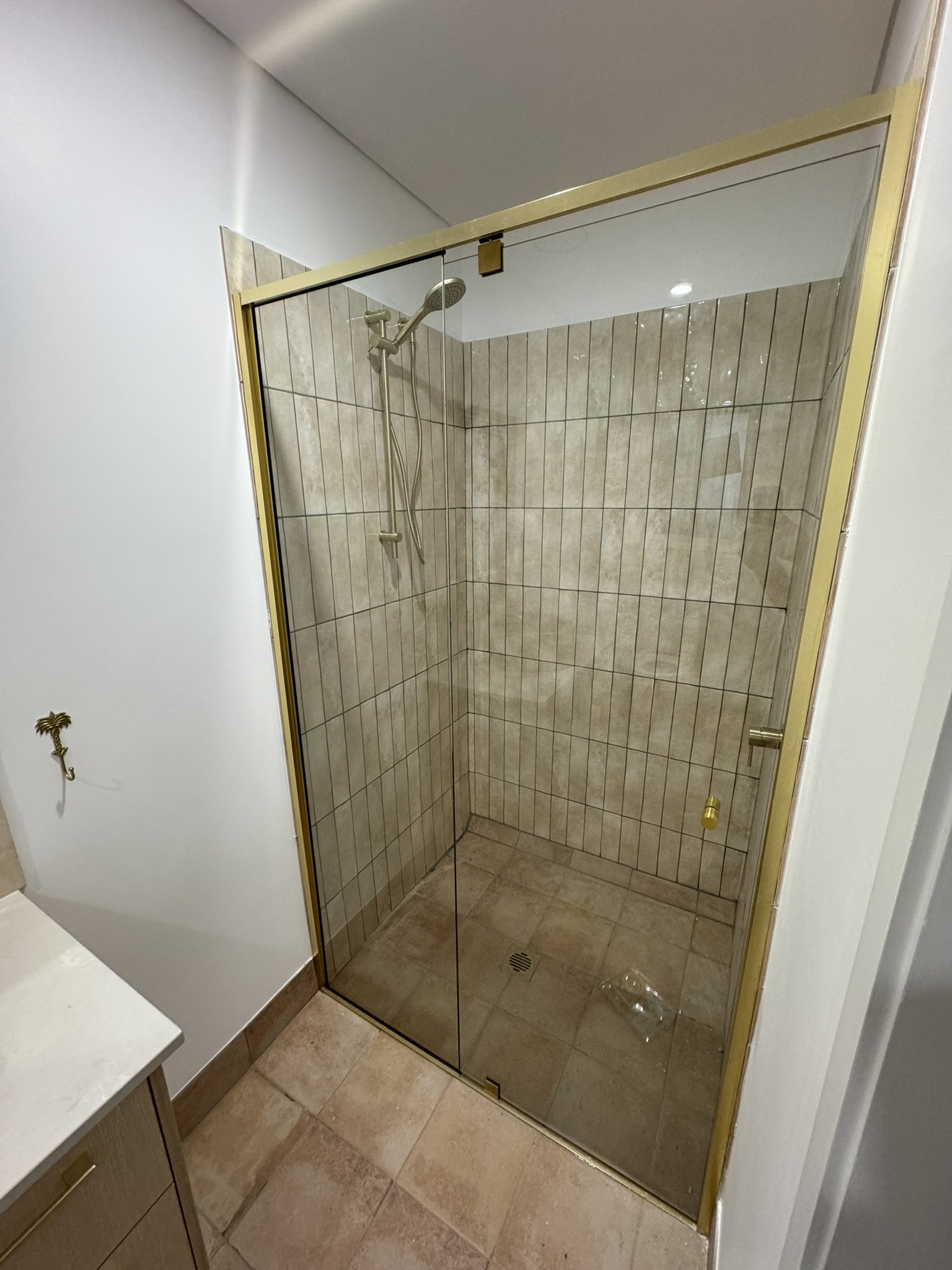 A walk-in shower with beige tiles and a glass door, featuring a handheld showerhead and a drain on the floor.