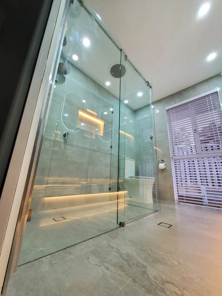 Modern glass-enclosed shower with multiple overhead and side LED lights, beige tiled walls, a window with blinds, and a floor drain.