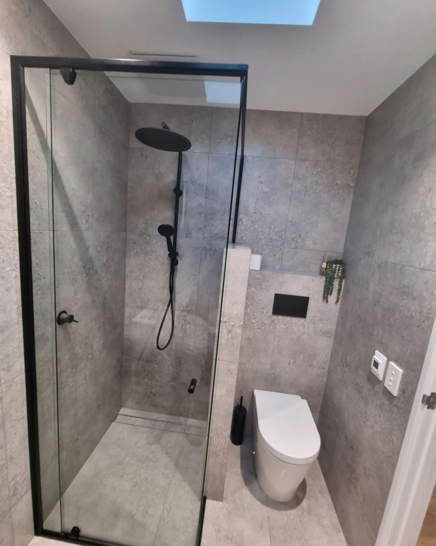 Modern bathroom with gray tiled walls and floor, glass shower enclosure with black frame, black handheld showerhead, white toilet, small wall-mounted shelf with green plant, toilet brush, and two hookups on the wall.