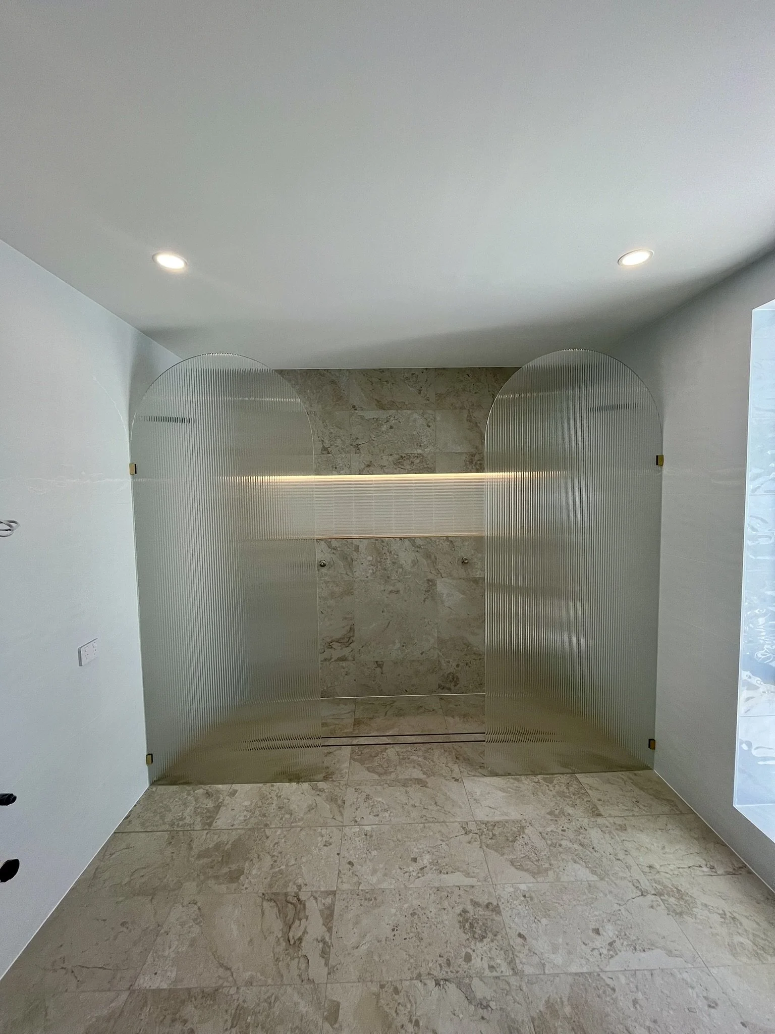 Interior of a modern room with beige marble flooring, light-colored walls, and a light gray ceiling with recessed lighting. There are two curved metallic or acrylic partitions with ridges in the center, and a tiled wall in the background.