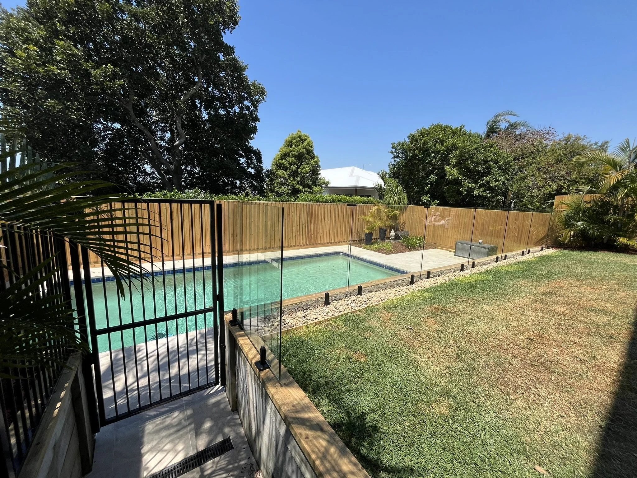 Backyard view featuring a swimming pool enclosed by a glass fence, green lawn, trees, and a wooden fence under a clear blue sky.