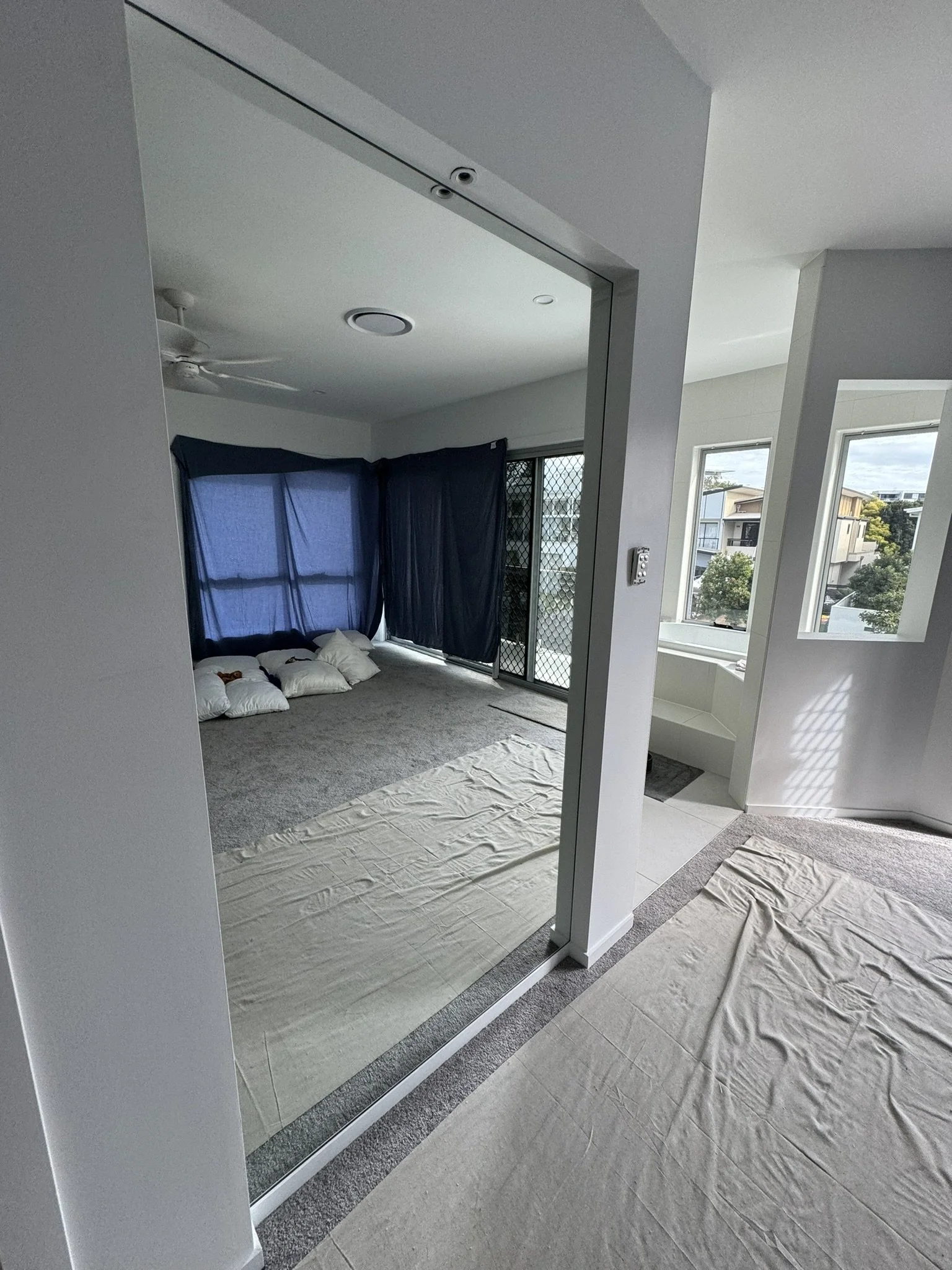 Interior of a modern apartment with a large mirror at the entrance, showing a bedroom with dark curtains, a ceiling fan, and windows with a view of neighboring buildings and trees.