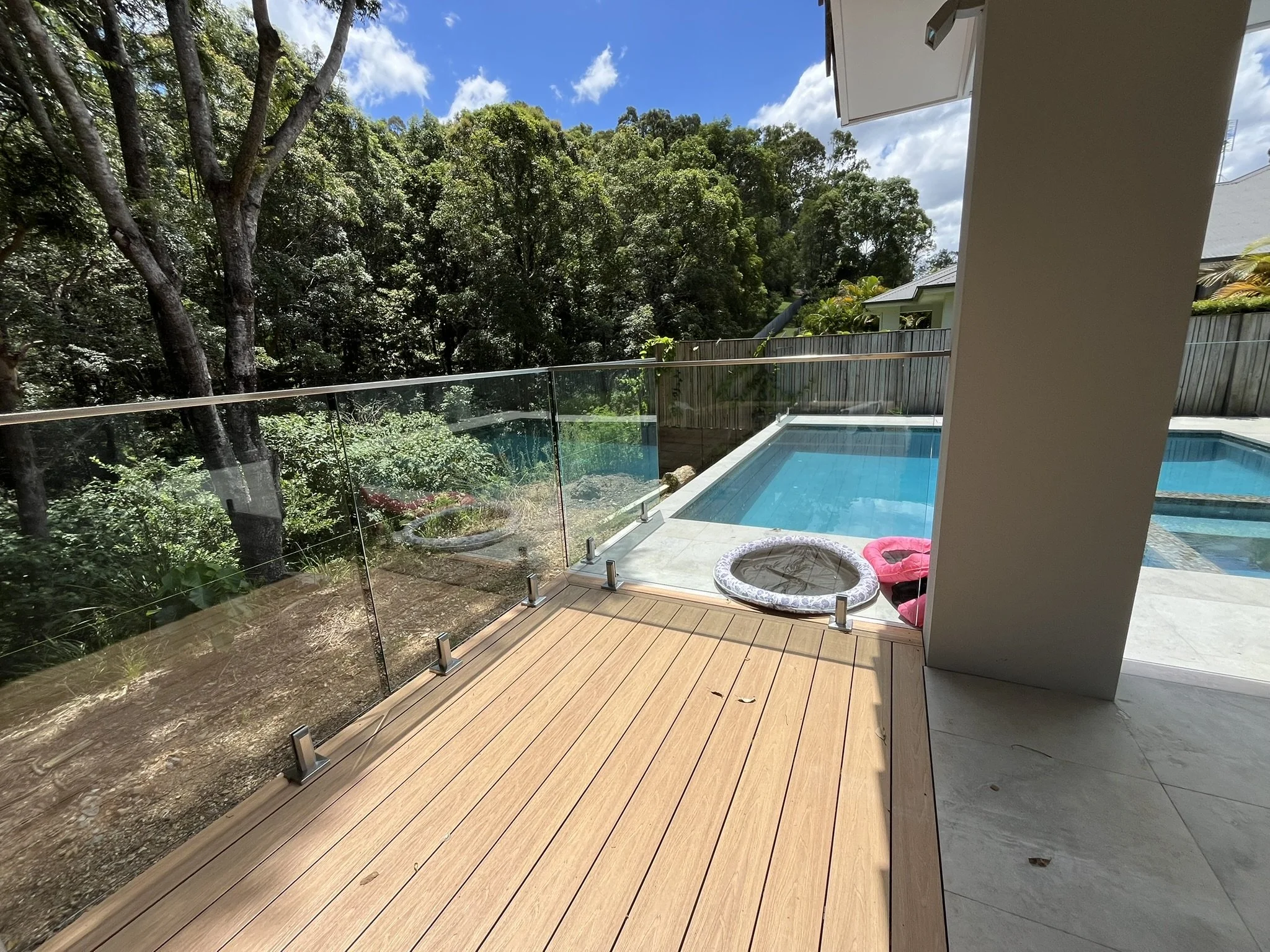 Balcony with wooden flooring, glass railing, a small pool or hot tub, and seating area, overlooking trees and a bright blue sky with clouds.