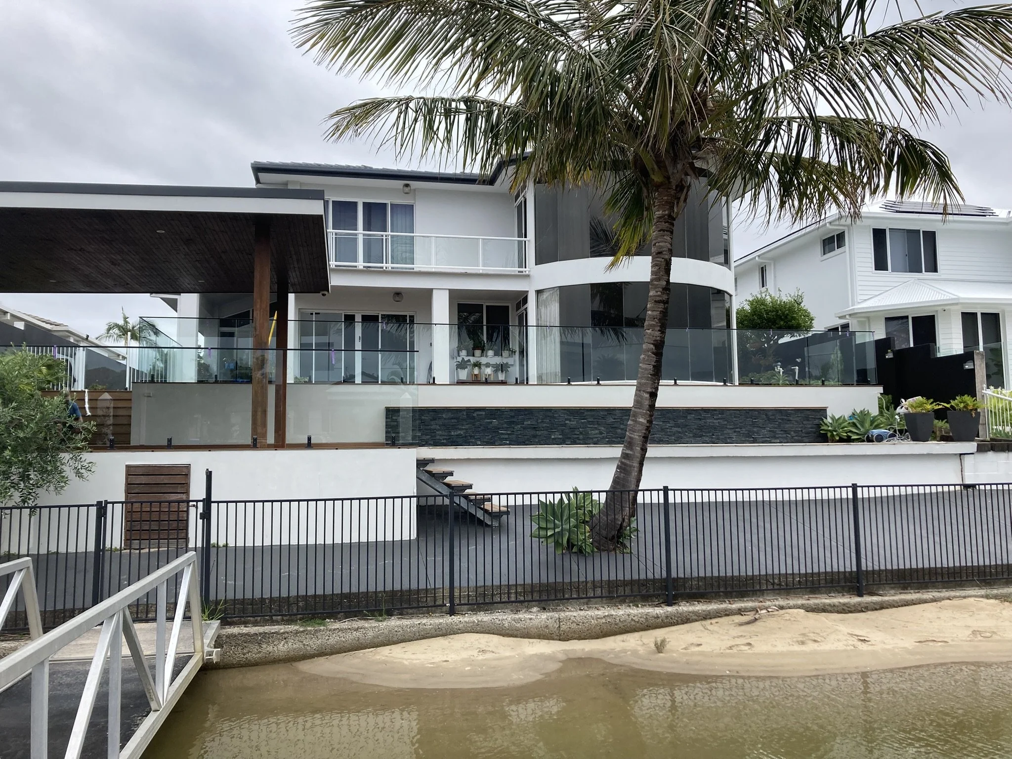 Modern two-story house with large windows, white exterior, balcony, palm tree in front, and a yard with a sandy area and water in the foreground.