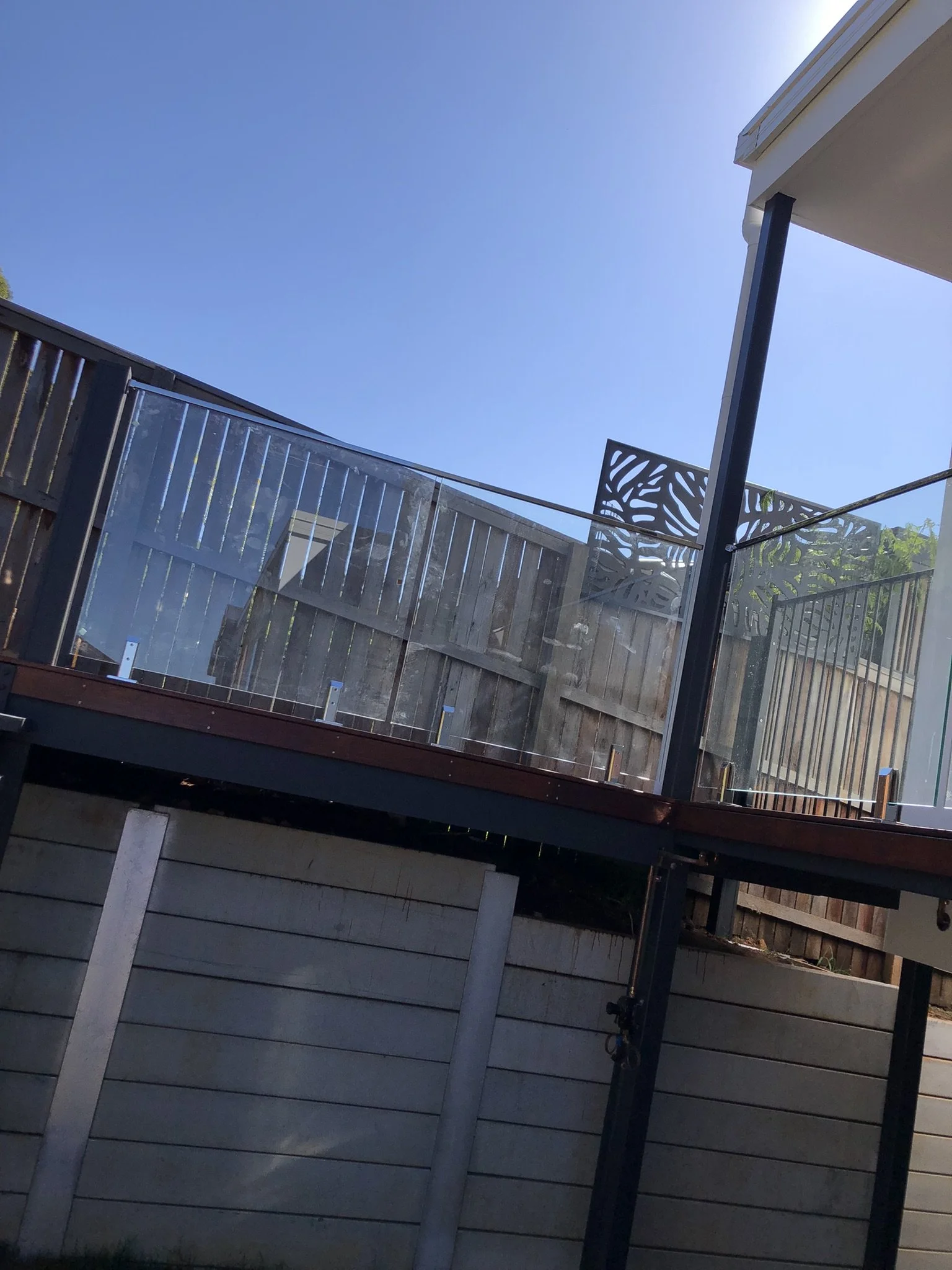 Photograph of a backyard patio with wooden fencing, a glass railing, and a decorative metal panel with leaf patterns, taken during daytime with clear blue sky.