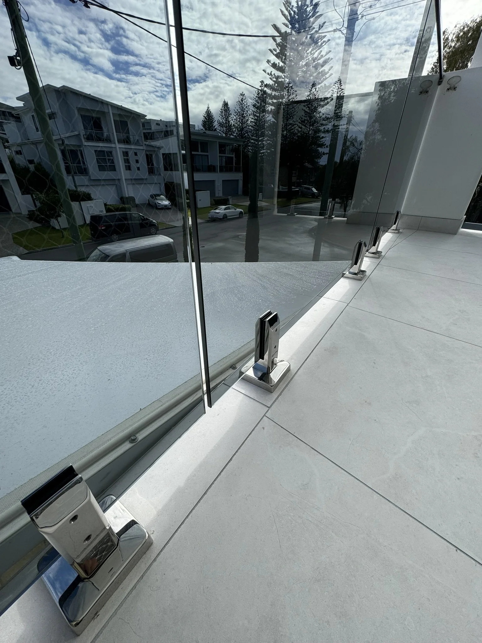 Glass doors with metal hardware on a modern building, reflecting parked cars, buildings, trees, and a cloudy sky outside.
