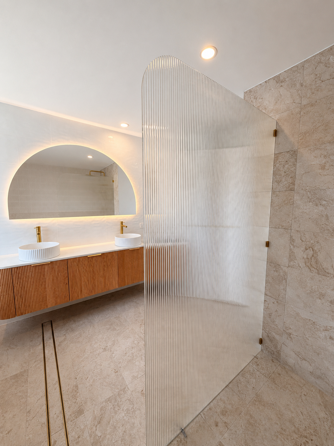 Modern bathroom with double vessel sinks, large mirror with backlighting, wooden floating vanity, textured glass privacy partition, beige tiled walls and floor, ceiling lights.