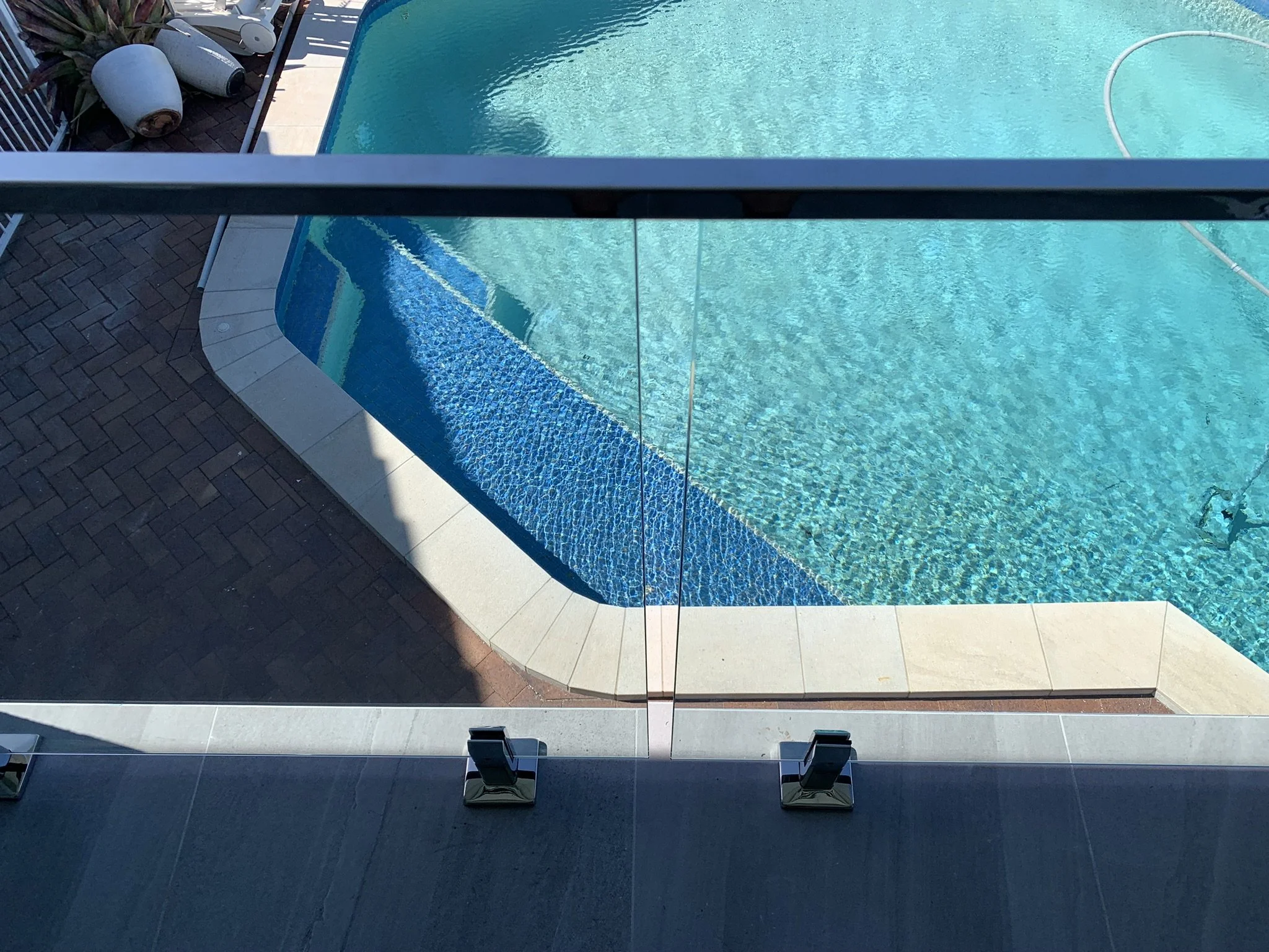 View of a swimming pool with clear blue water, a curved edge with mosaic tile detailing, and surrounding patio area with some potted plants and a terrace railing.