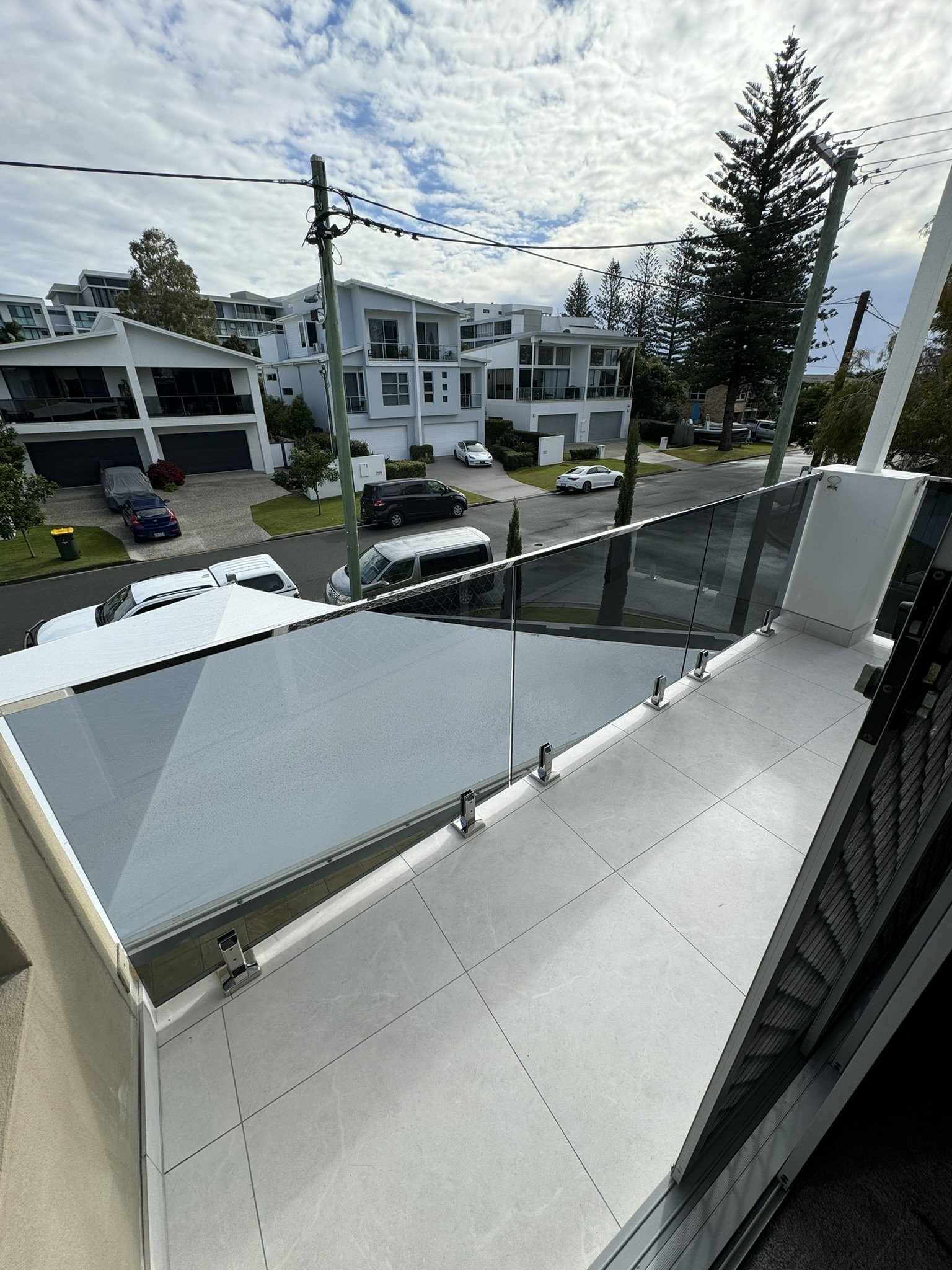 Modern balcony with glass railing overlooking a residential street with parking and contemporary white houses, cloudy sky, and trees.