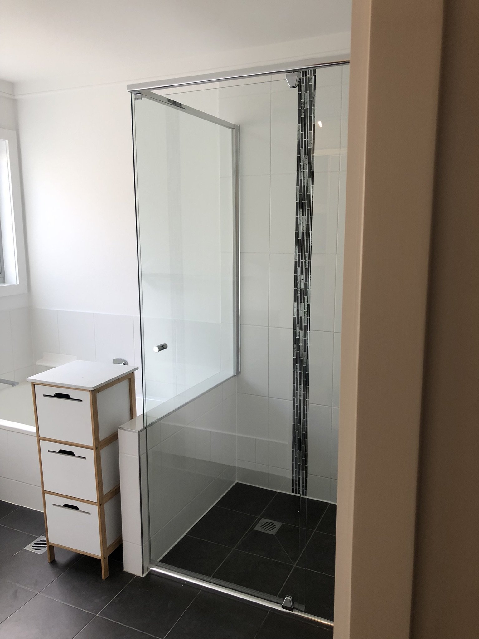 A modern bathroom with a glass shower enclosure, white tile walls, and a vertical strip of decorative black, gray, and silver tile along the shower wall. There is a small wooden storage unit with three drawers beside the shower, and a window on the l