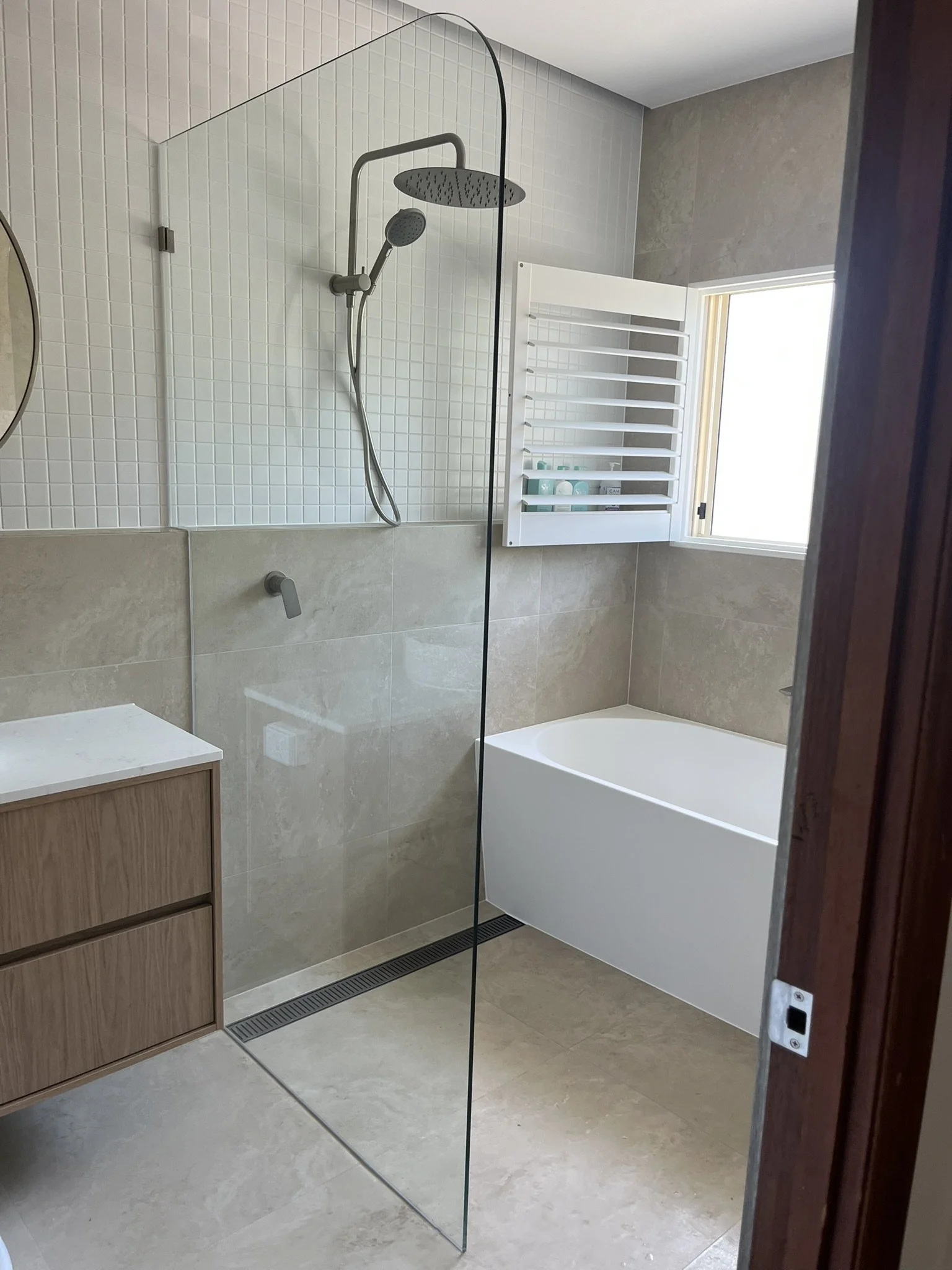 Modern bathroom with glass shower partition, white tiled wall, beige tiled floor, wall-mounted shower fixtures, a bathtub, and a small window.