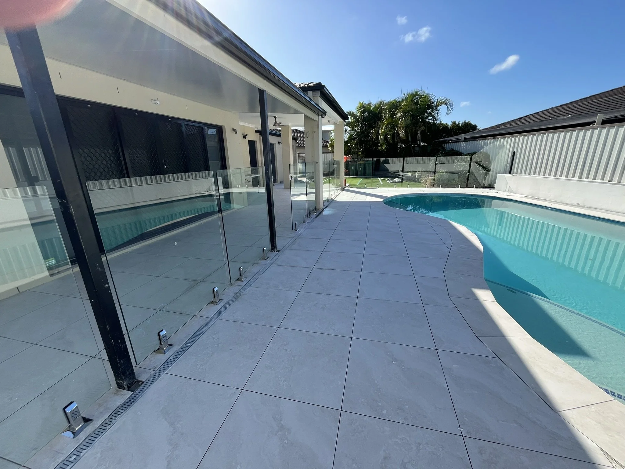 Backyard with swimming pool, tiled patio, glass pool fencing, house exterior, palm trees, and clear blue sky.