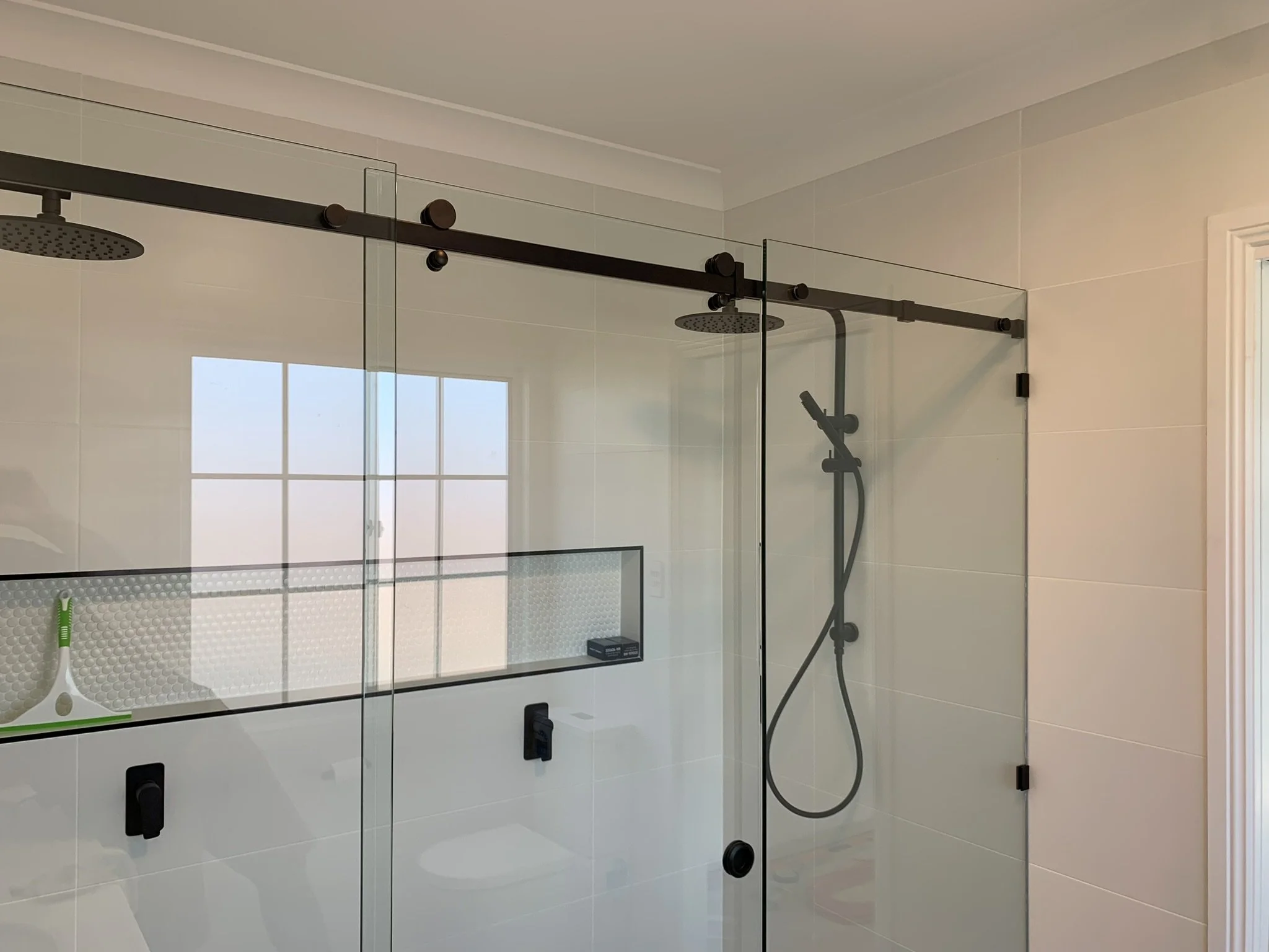 Glass-enclosed shower with two rainfall showerheads on a black metal rail, one handheld shower, a white tiled wall, a window with a grid pattern, and a small black object on the shower floor.