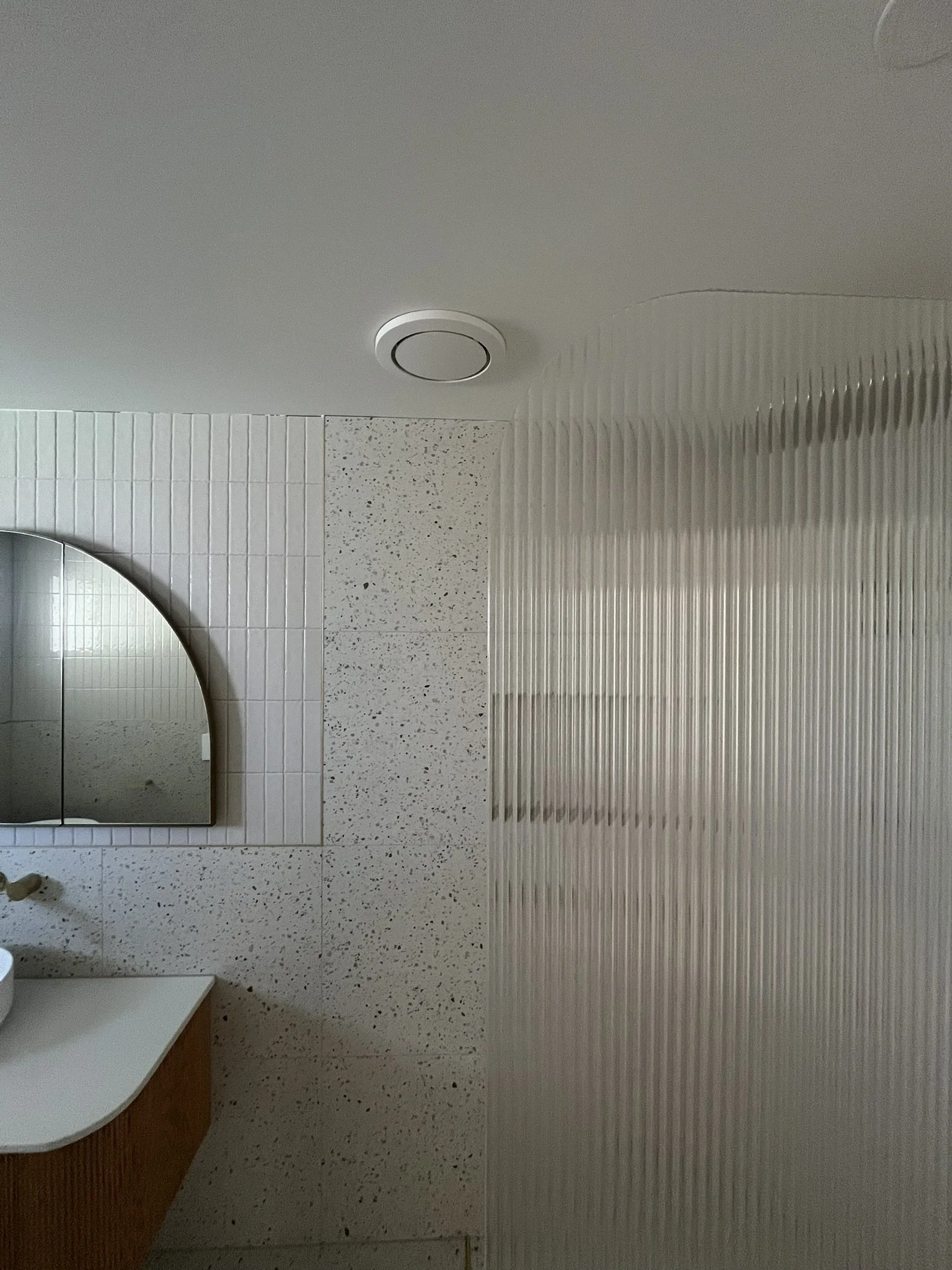Interior view of a bathroom ceiling with a round smoke detector, part of a mirror, and tiled walls with a textured panel.