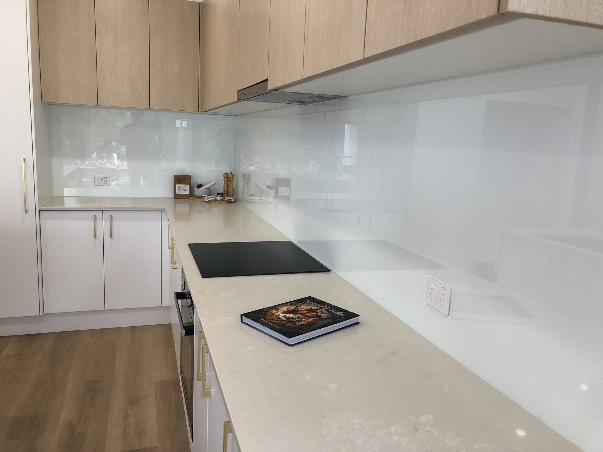 A modern kitchen with light wood cabinets, a white countertop, and a glossy white backsplash. There are a few decorative items and a large book on the counter.