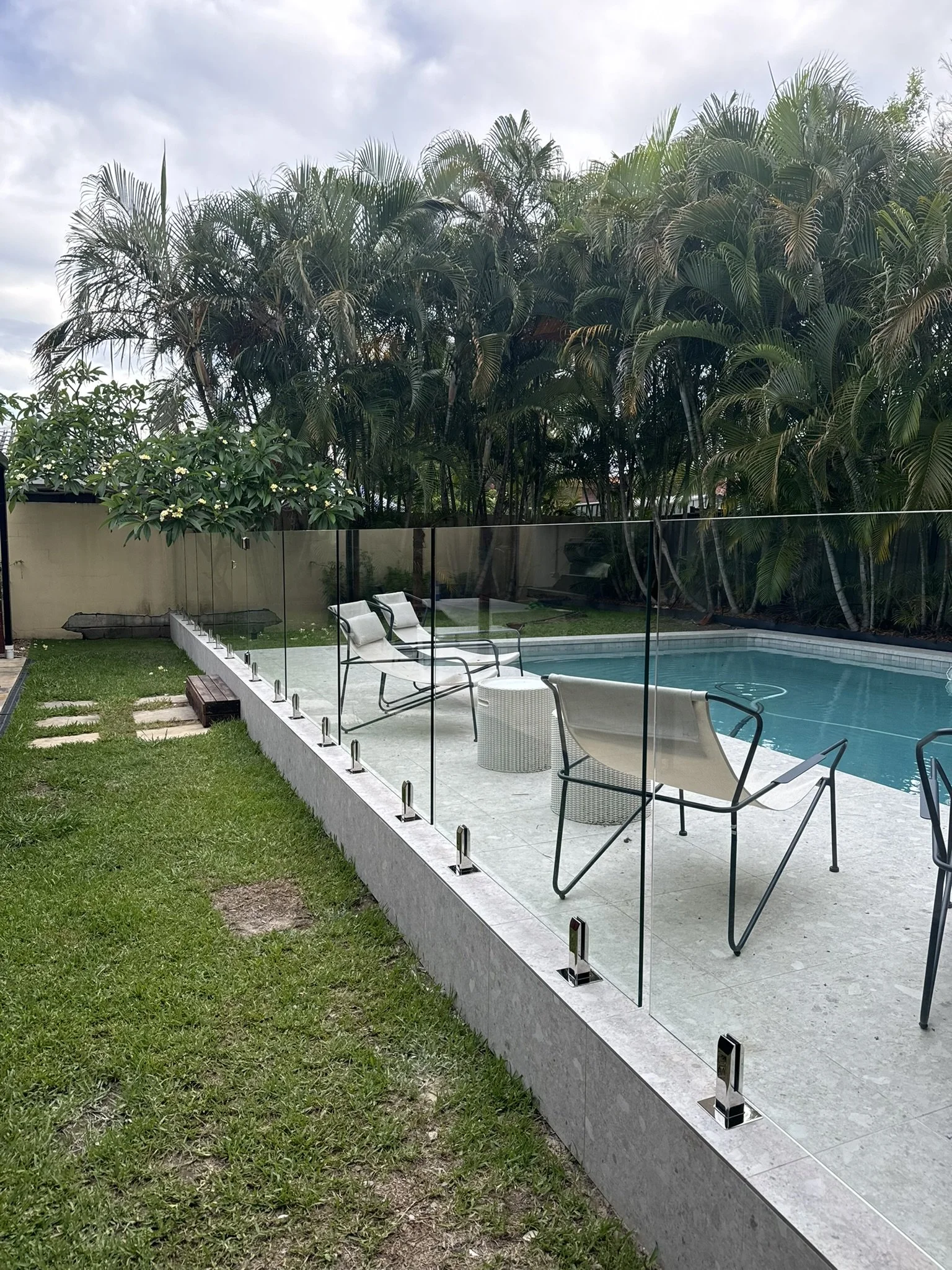Backyard with a swimming pool, glass fence, lounge chairs, green grass, and tall palm trees under cloudy sky.