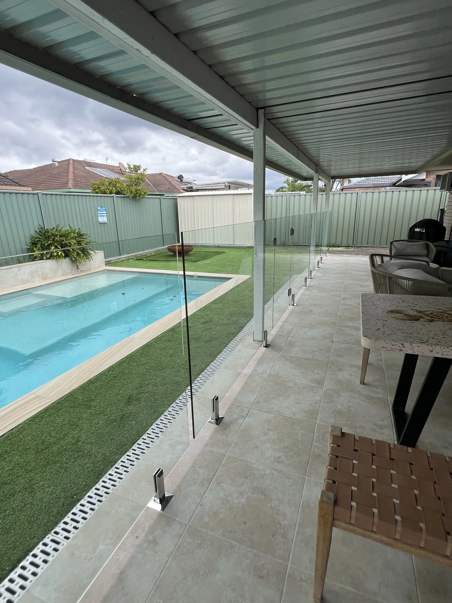 View of a backyard patio area with a swimming pool, artificial grass, and a glass safety barrier. There are some outdoor chairs and a table on a tiled floor, enclosed by a green metal fence with neighboring rooftops and cloudy sky overhead.