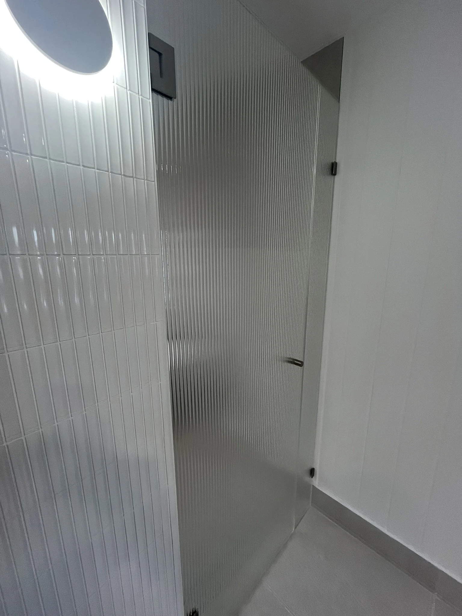 An indoor space with white tiled walls and light grey floor tiles. There are two metal doors with vertical grooves and black handles, one in the foreground and one further back, creating a corridor or room divider effect.