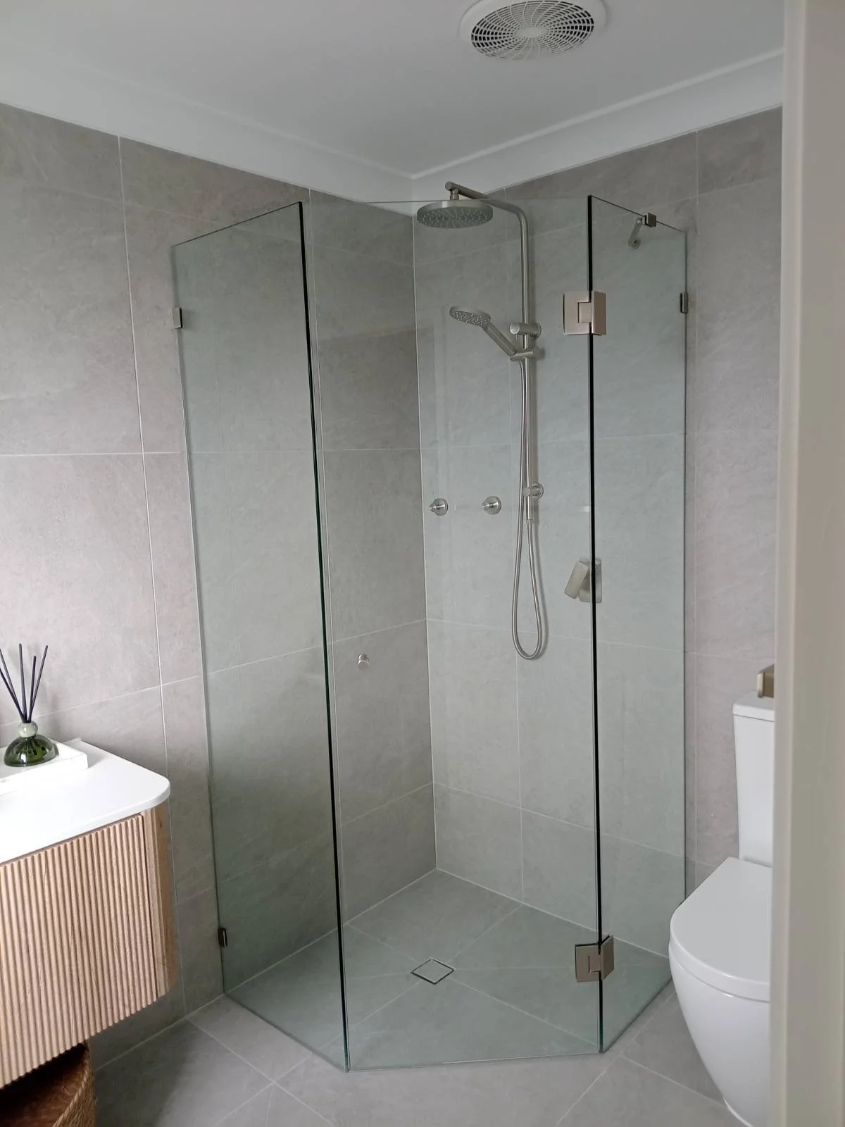 A modern bathroom with a glass shower enclosure, grey tiled walls and floor, a ceiling vent, a rainfall shower head, a handheld shower wand, a white toilet, and a small cabinet with a diffuser on top.