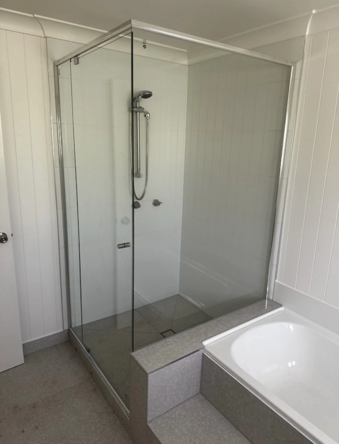 A bathroom shower area enclosed with glass panels, with a handheld showerhead and wall-mounted controls, next to a built-in bathtub.