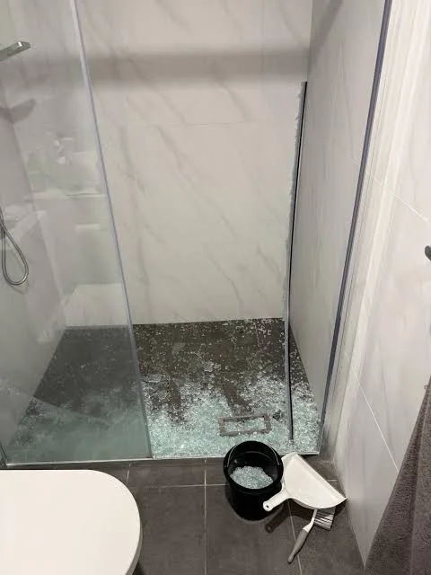 Shower area with broken glass, a black bucket, a white scraper, and a lint roller on the bathroom floor.