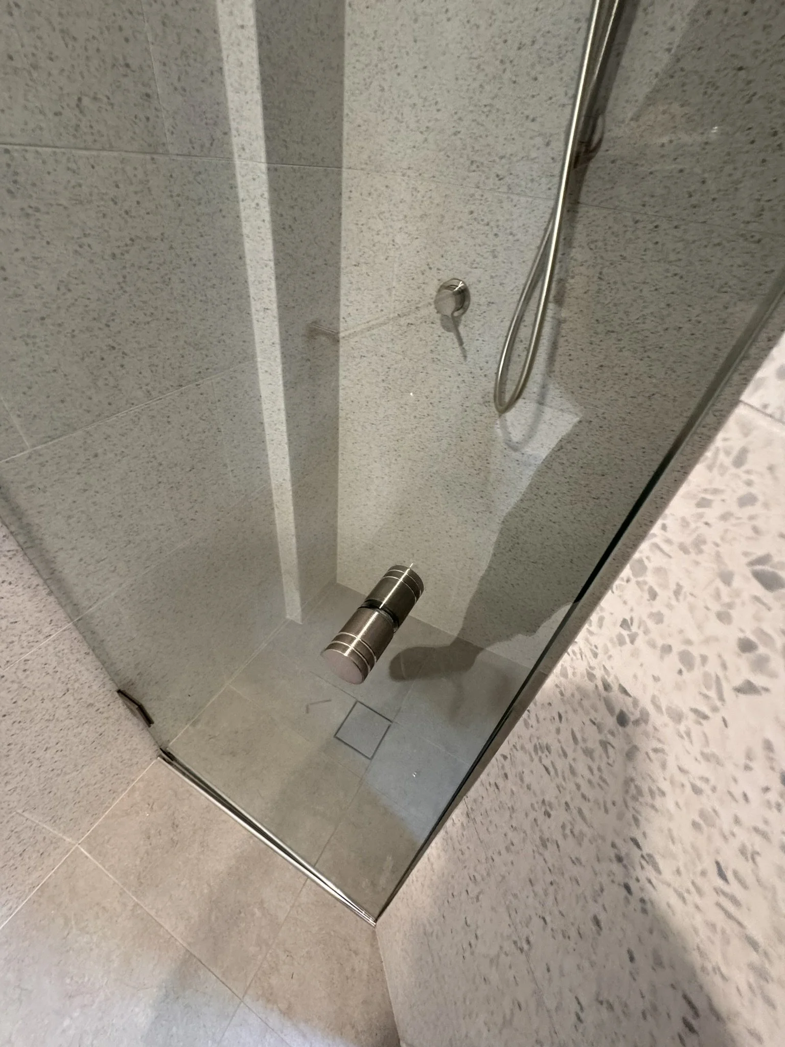 A bathroom shower area with a gray tiled wall, metal handle, and a shower head with a flexible hose. There is a glass shower door with a metal hinge.