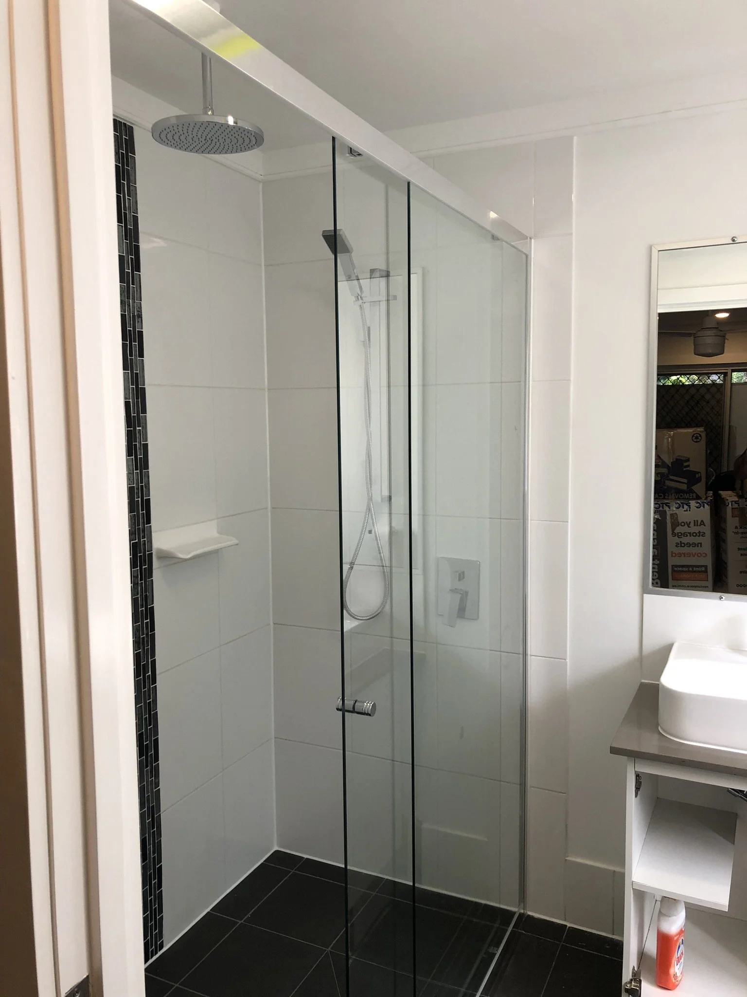 A modern bathroom with a glass shower enclosure, white tiled walls, a black strip of small tiles, a showerhead, a handheld shower, a small white shelf inside the shower, and a white sink with a cabinet. There is a mirror on the right side and a fire 