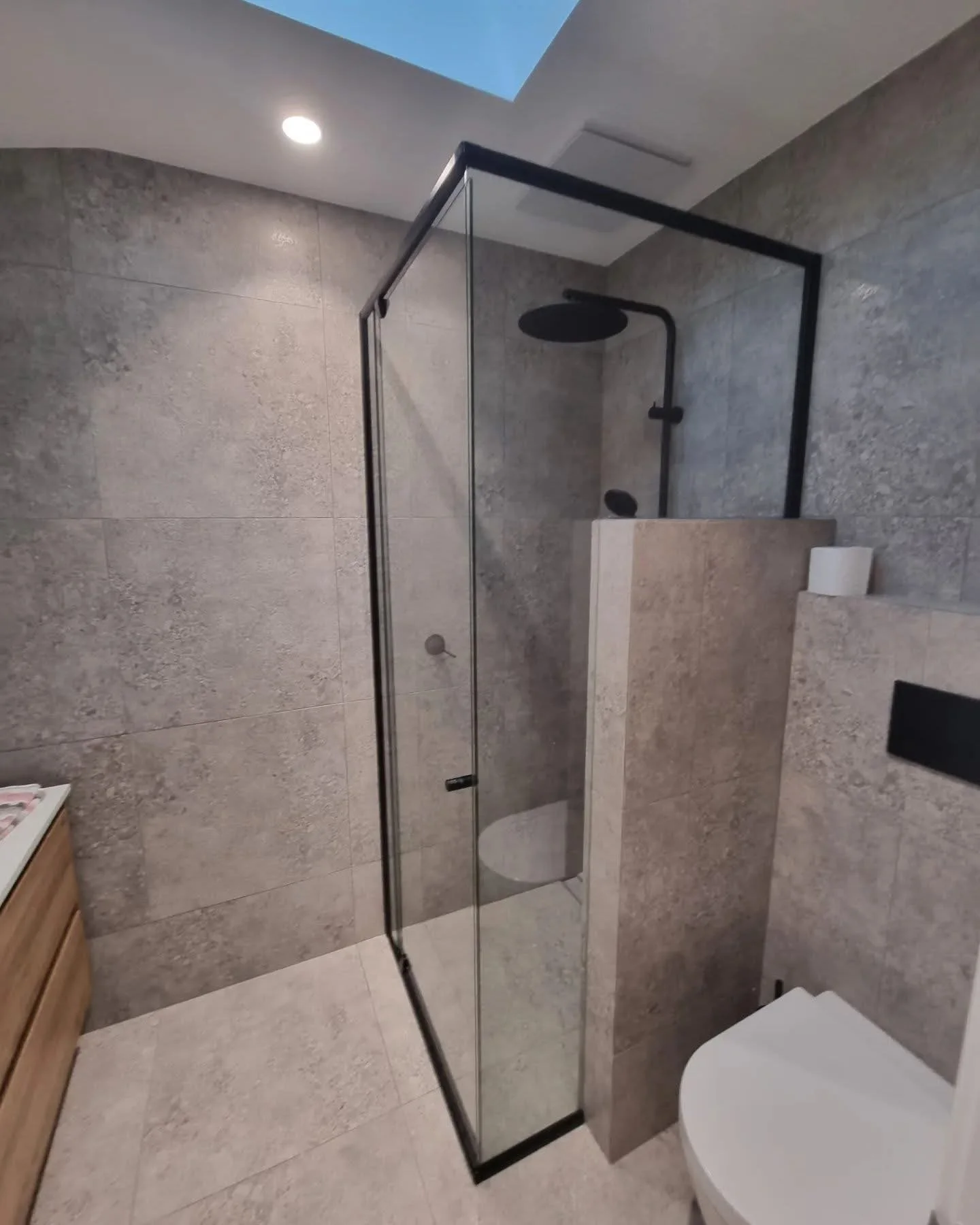 Modern bathroom with gray textured tile walls and floor, glass shower enclosure with black frame and black shower fixtures, and a white toilet visible in the corner.