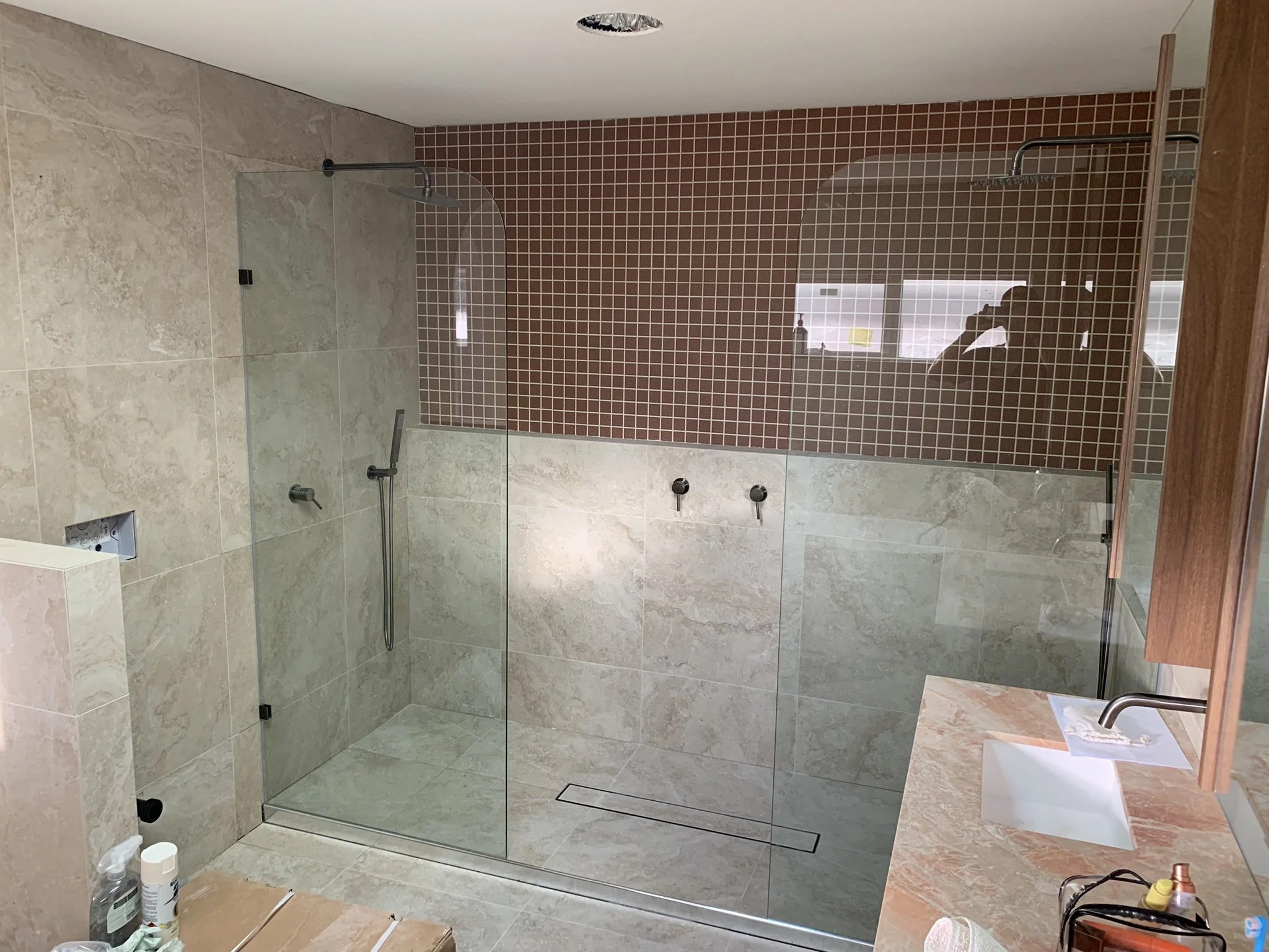 A modern bathroom with a large walk-in shower featuring clear glass walls, a rain shower head on a red mosaic tile wall, beige marble-like tiles on the other walls, a wooden vanity with a sink, and various toiletries on the counter.