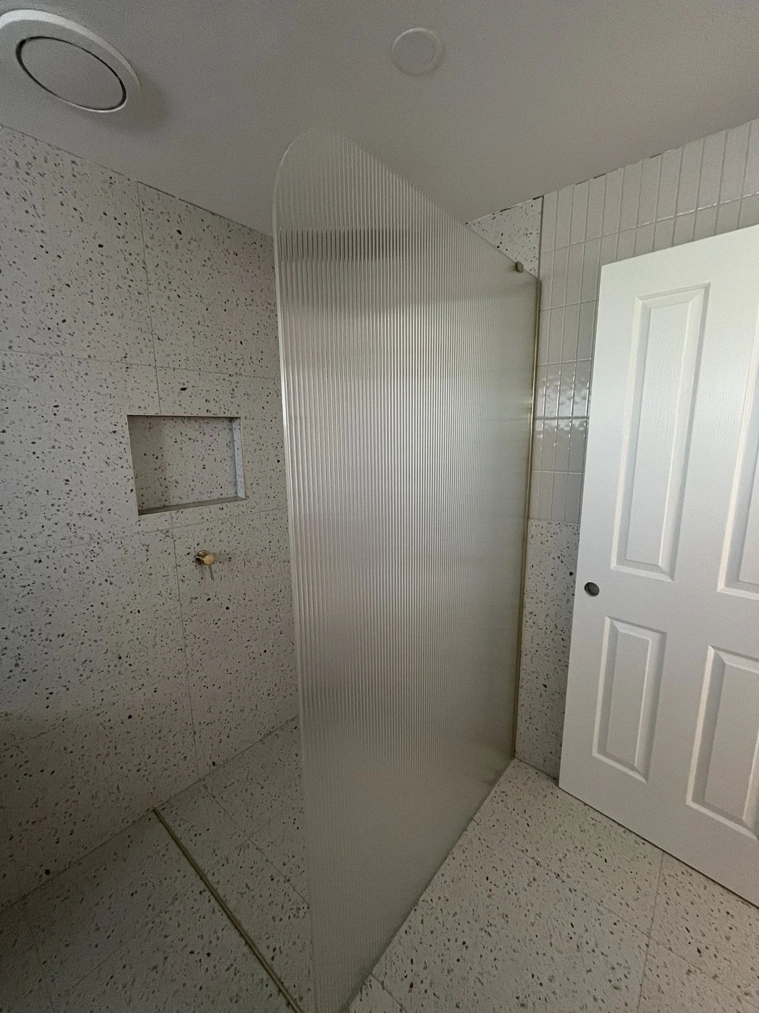 A bathroom with speckled tile floor and walls, a white door leaning against the wall, a textured shower partition, and a built-in shelf or niche in the wall.