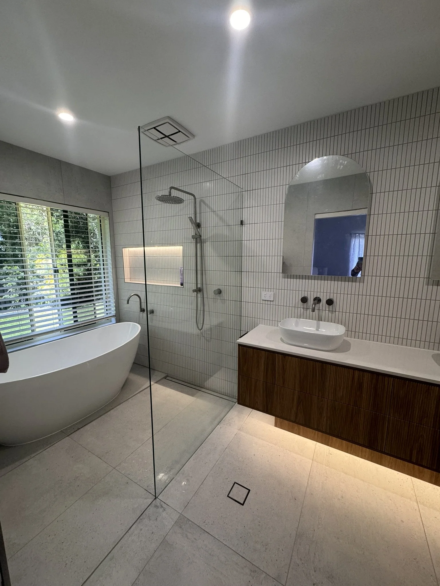 Modern bathroom with a freestanding bathtub, glass shower enclosure, wall mirror, wooden vanity with vessel sink, tiled wall, and large window with greenery outside.