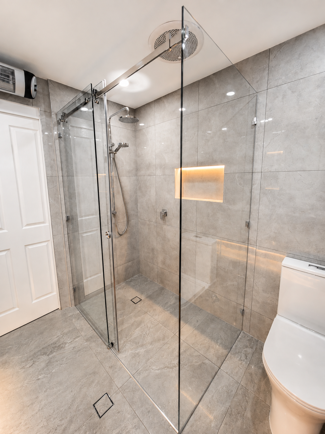 Modern walk-in shower with glass enclosure, multiple showerheads, and wall niche for toiletries, in a bathroom with beige tiles.