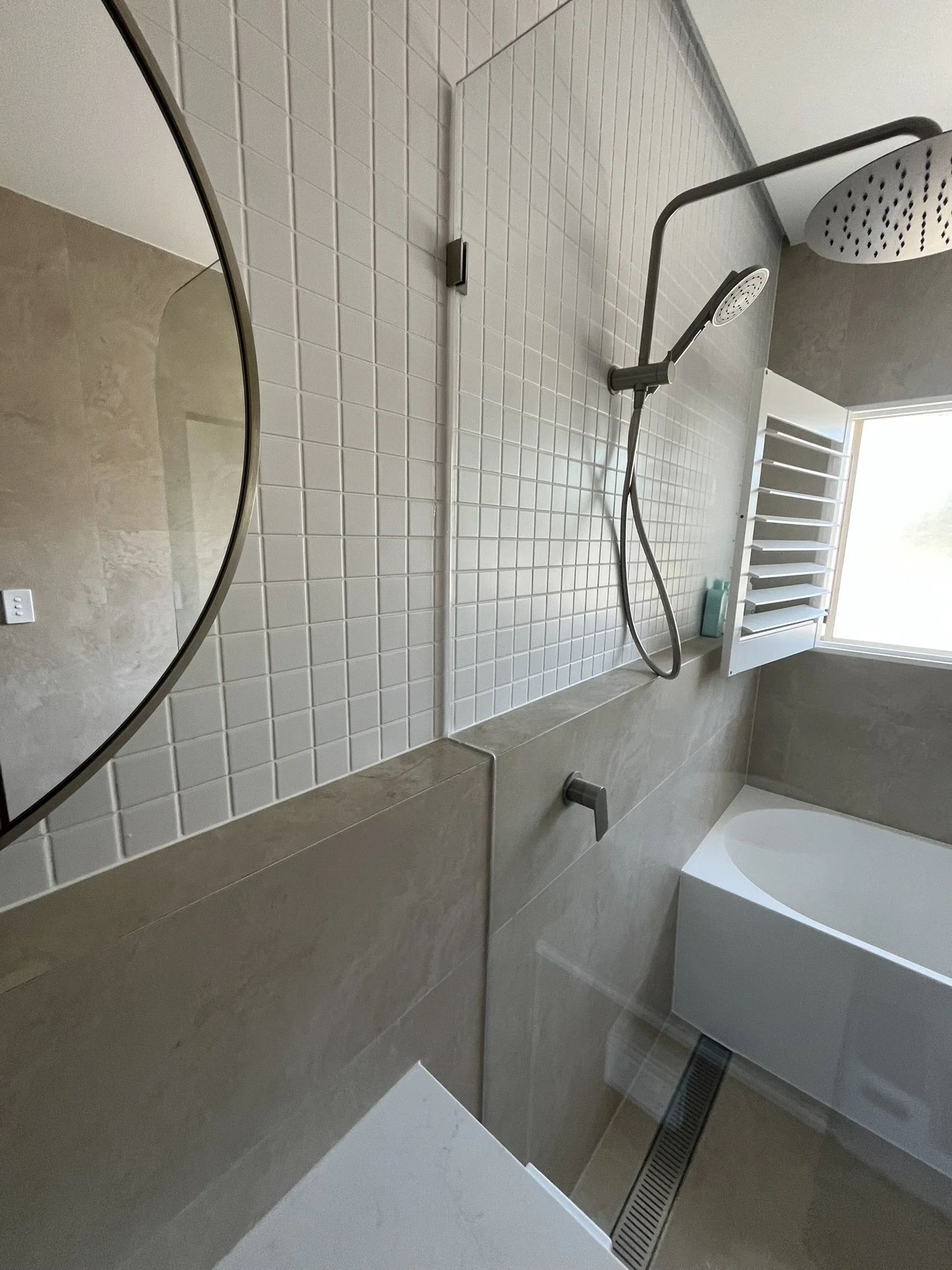 Modern bathroom shower area with white square tiles, a black handheld showerhead, a white bathtub, a small mirror on the wall, a window letting in natural light, and a light gray floor.