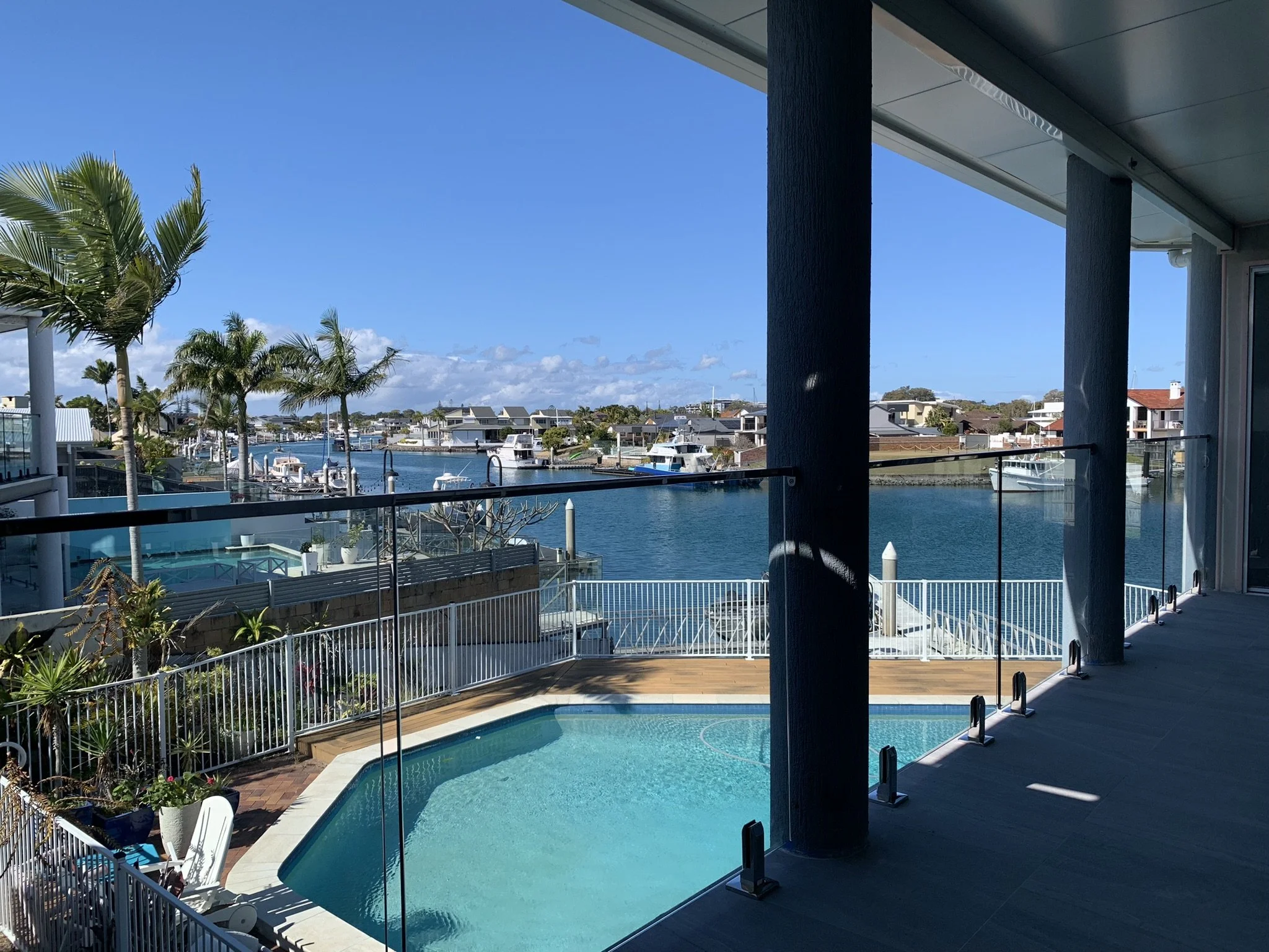 View of a marina with boats, surrounded by houses, palm trees, and a clear blue sky, from a balcony with a swimming pool in the foreground.