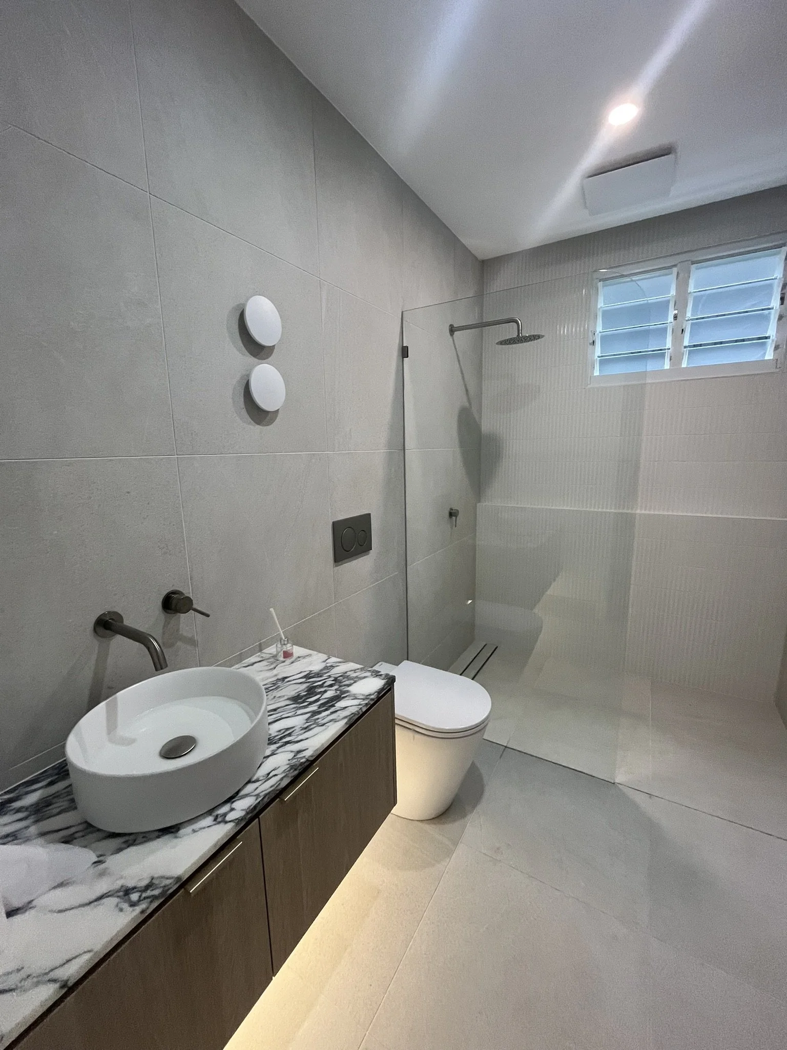 A modern bathroom with a marble countertop, vessel sink, toilet, walk-in shower, window with blinds, toilet paper holder, and recessed lighting.