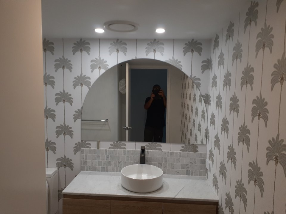 Bathroom with wallpaper featuring palm tree patterns, a large mirror, a white vessel sink on a marble countertop, and a person taking a photo visible in the mirror.