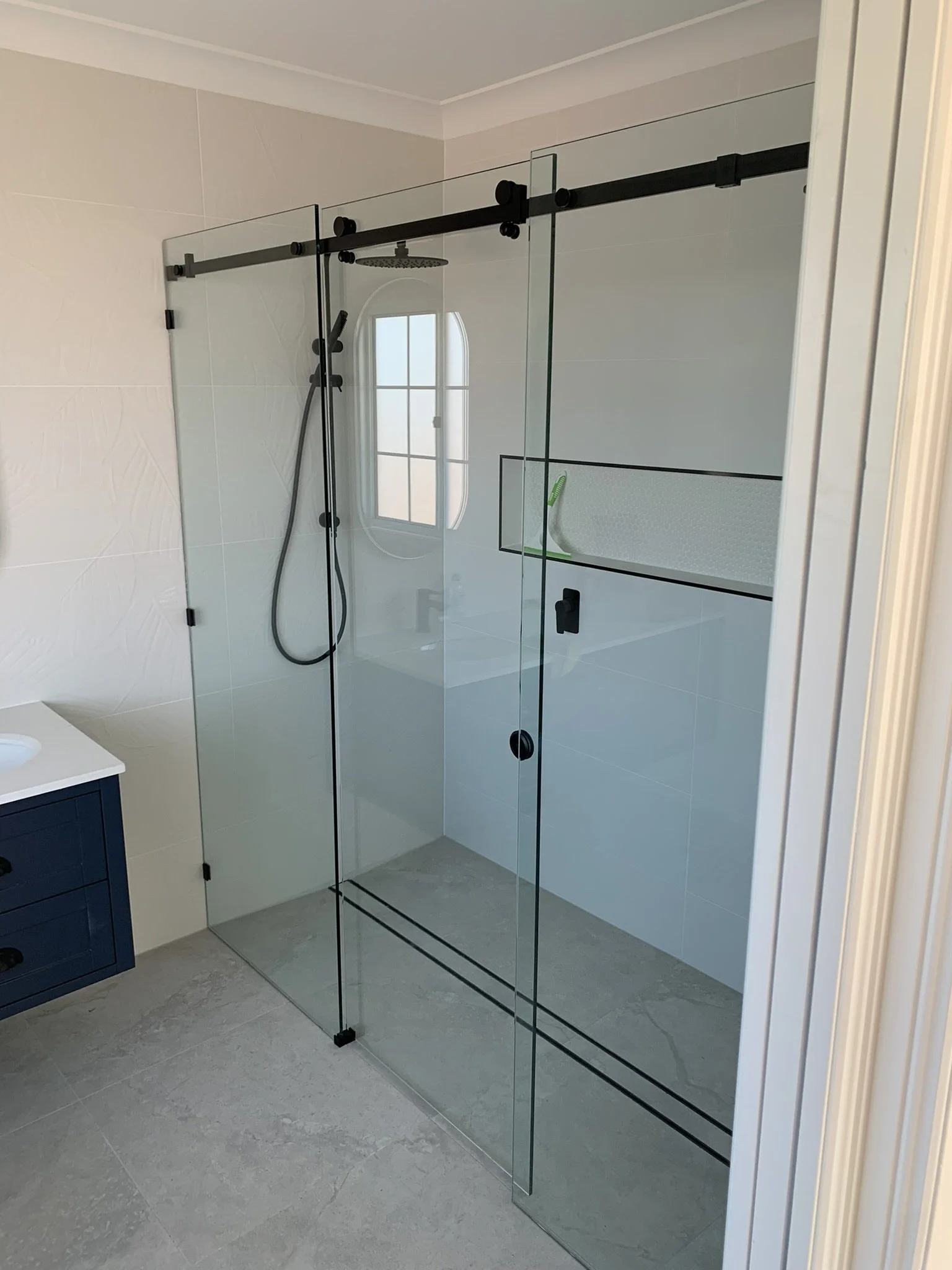 A modern glass shower stall with a black sliding barn door hardware system, a rain showerhead, a handheld showerhead, inside a bathroom with white tiled walls, a frosted window, and a blue bathroom vanity with a white countertop.