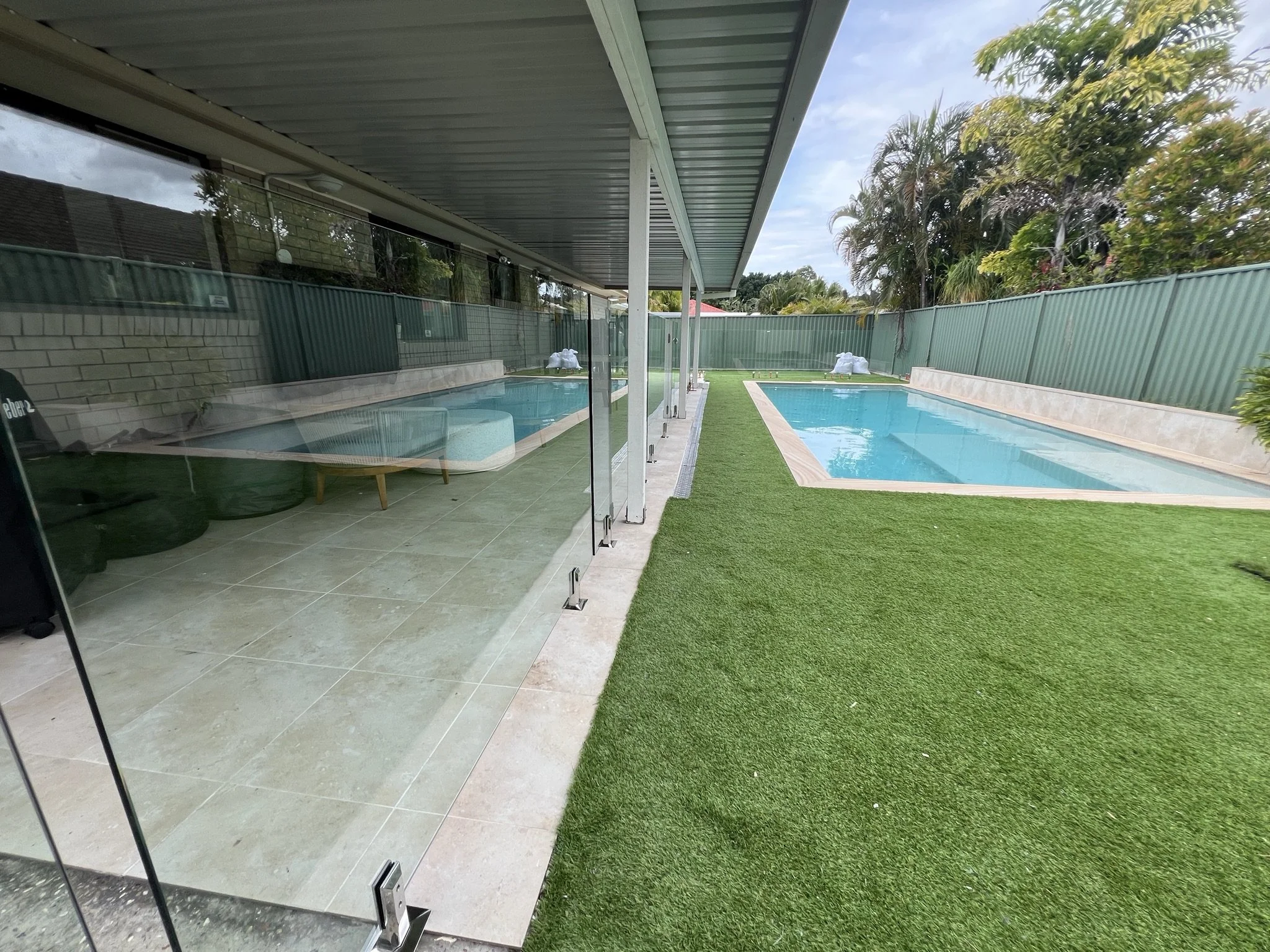 View of backyard pool area with a glass partition, a small lounge chair, green artificial grass, and lush trees in the background under a partly cloudy sky.