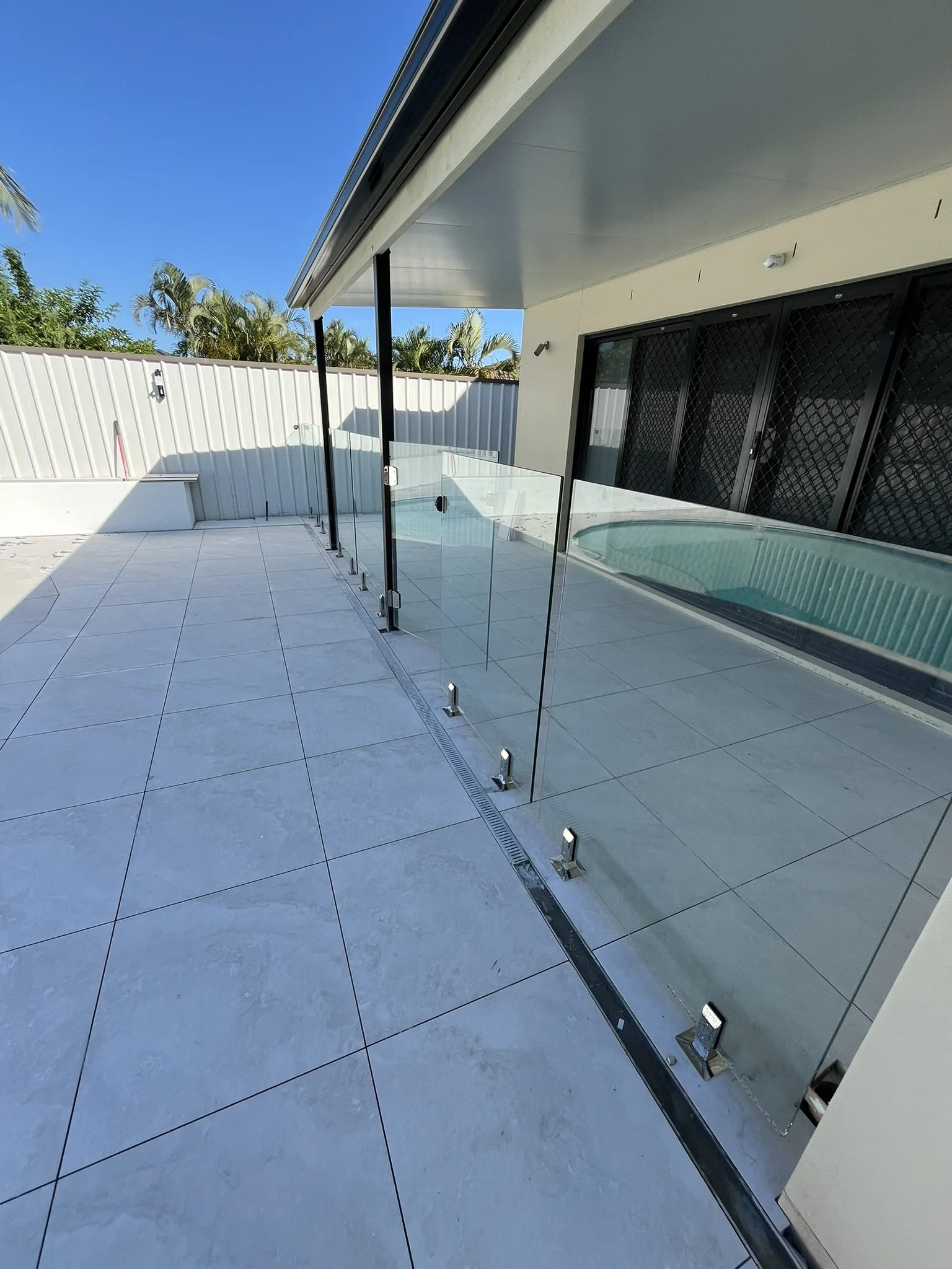 Backyard patio with tiled floor, glass pool fencing, and a hot tub under a covered area. Fence with palm trees visible in the background, and sliding door with security mesh.