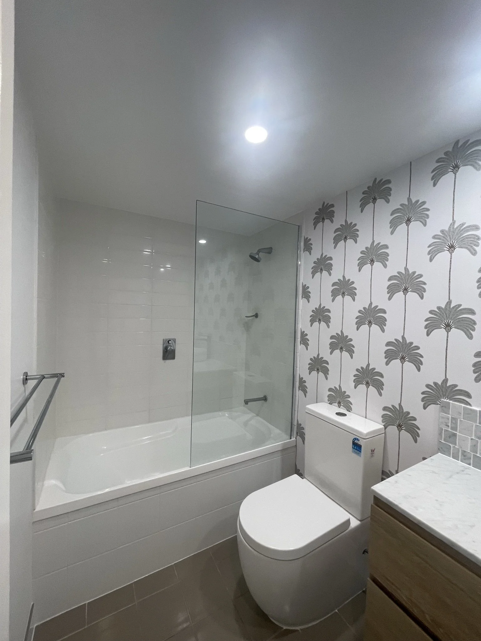 Modern bathroom with a white bathtub and shower, a glass shower door, a wall with palm tree wallpaper, a white toilet, and a marble countertop with a wooden vanity.