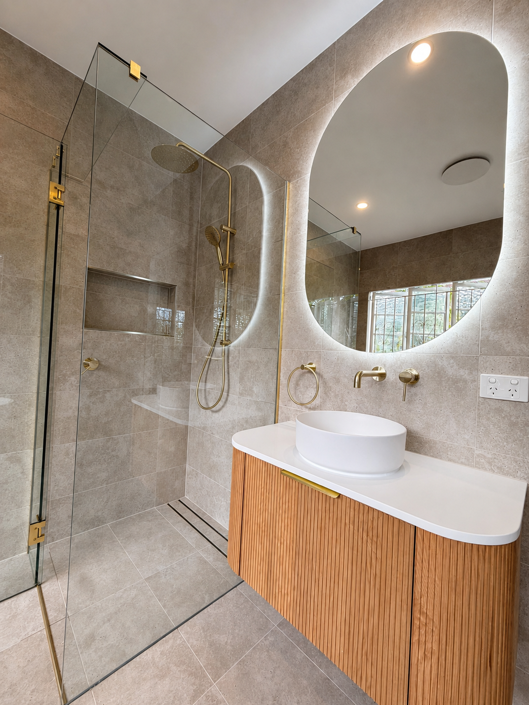 Modern bathroom with walk-in shower featuring gold fixtures, a large mirror, a white vessel sink on a wooden vanity, beige tiled walls and floor, and recessed ceiling lighting.