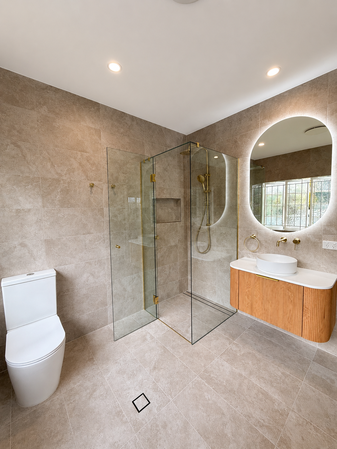 Modern bathroom with beige tiled walls and floor, a white toilet, a glass shower enclosure with gold fixtures, a wooden vanity with a round white vessel sink, an oval mirror, and a window with metal bars.
