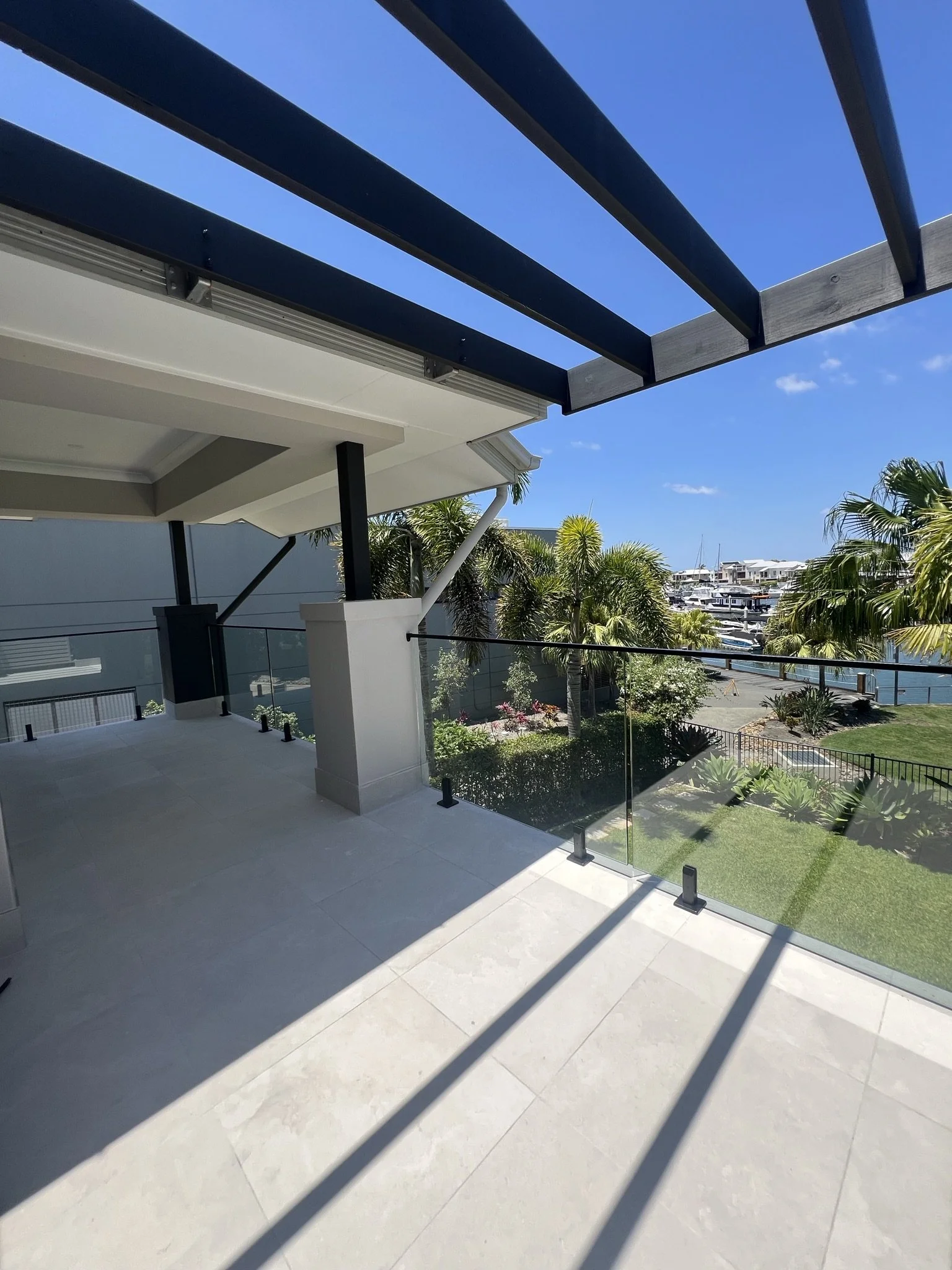 A balcony with glass railing, view of green trees, shrubs, and boats docked in a marina under a clear blue sky.