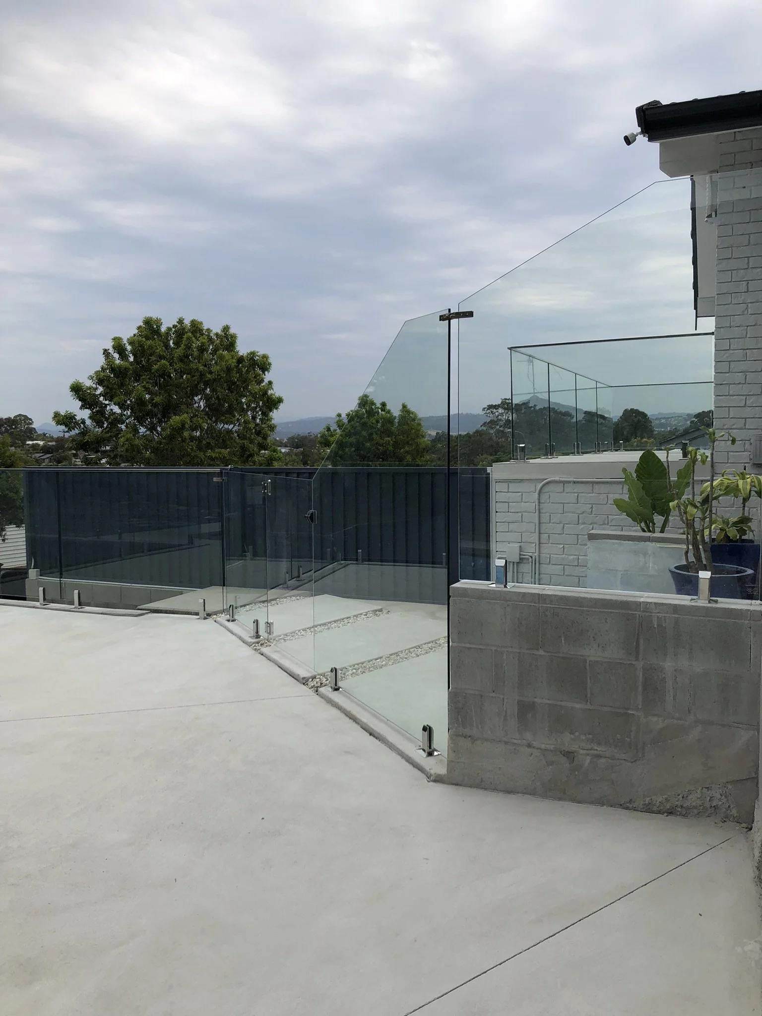 A modern outdoor balcony with glass safety railing, concrete flooring, the corner of a white brick building with a plant in a pot, and trees and cloudy sky in the background.