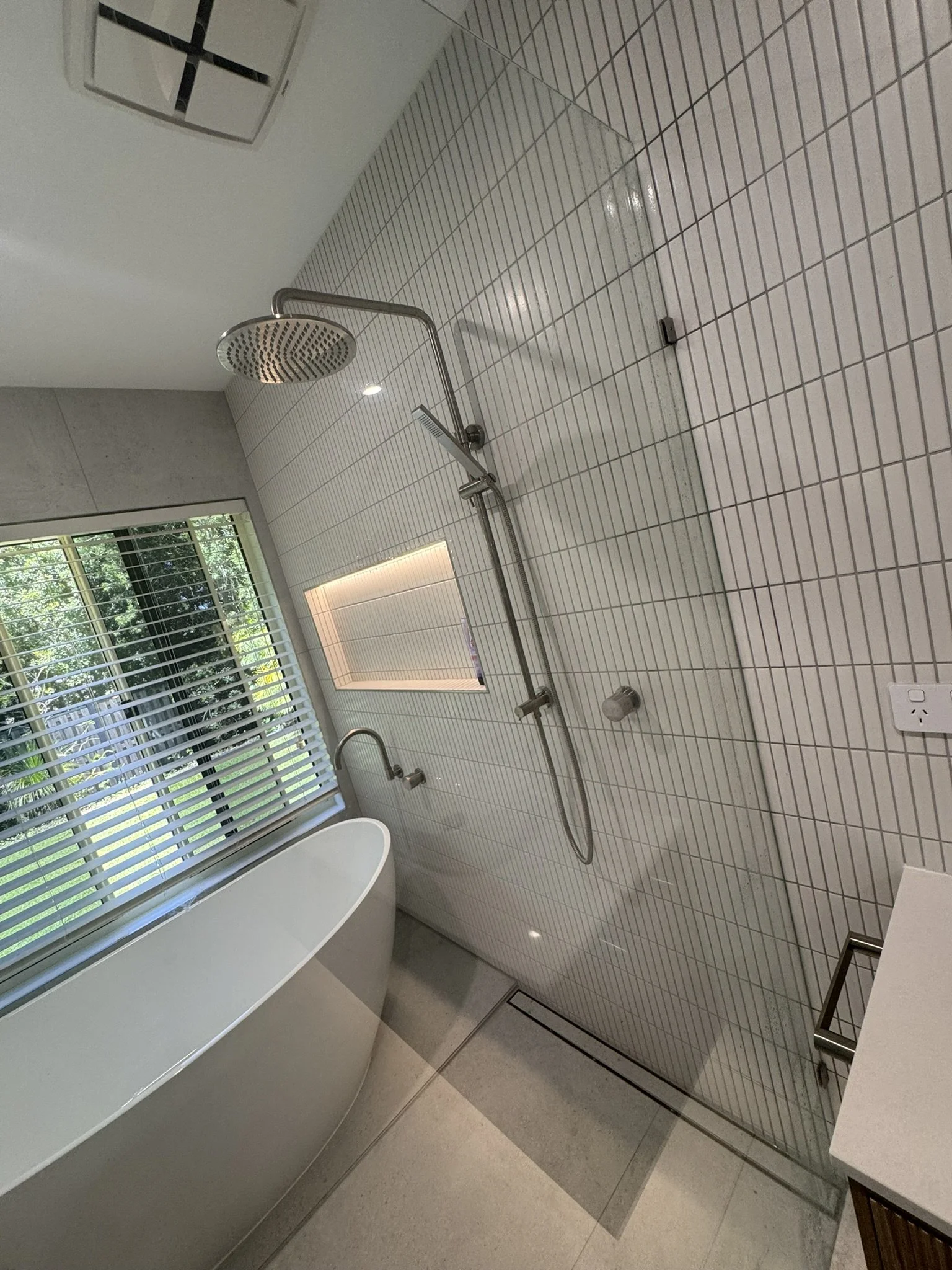 Modern bathroom with a freestanding bathtub, a rain showerhead, a window with blinds, and minimalist decor.