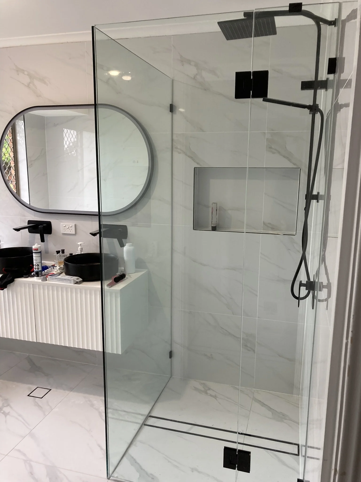 Modern bathroom with a glass shower enclosure, wall-mounted black showerhead, handheld shower, and built-in niche for toiletries. White marble tiles on walls and floor.