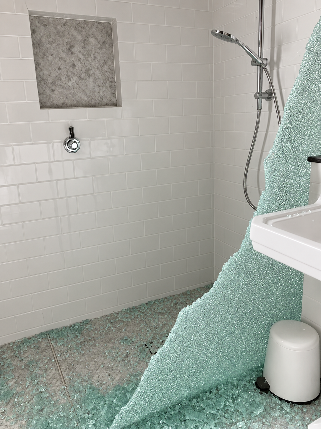 A bathroom with a large shower area that has very broken glass on the floor and folded broken glass on the corner of a sink and trash can.