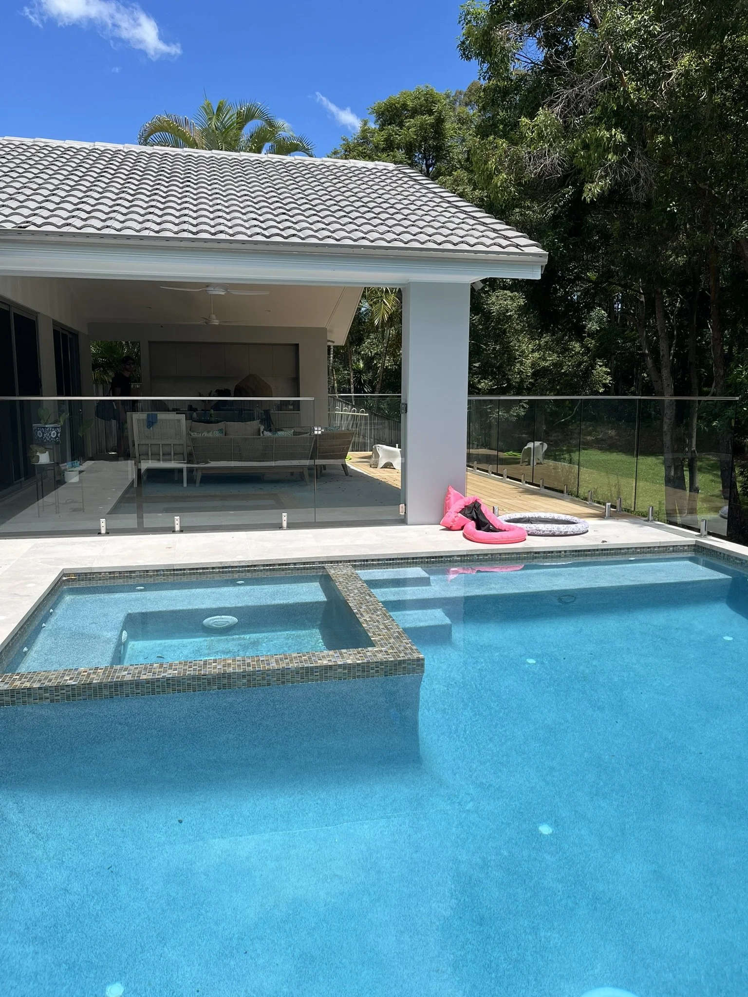 View of a backyard with a swimming pool in the foreground, a modern house with a covered patio, and lush trees in the background under a clear blue sky.