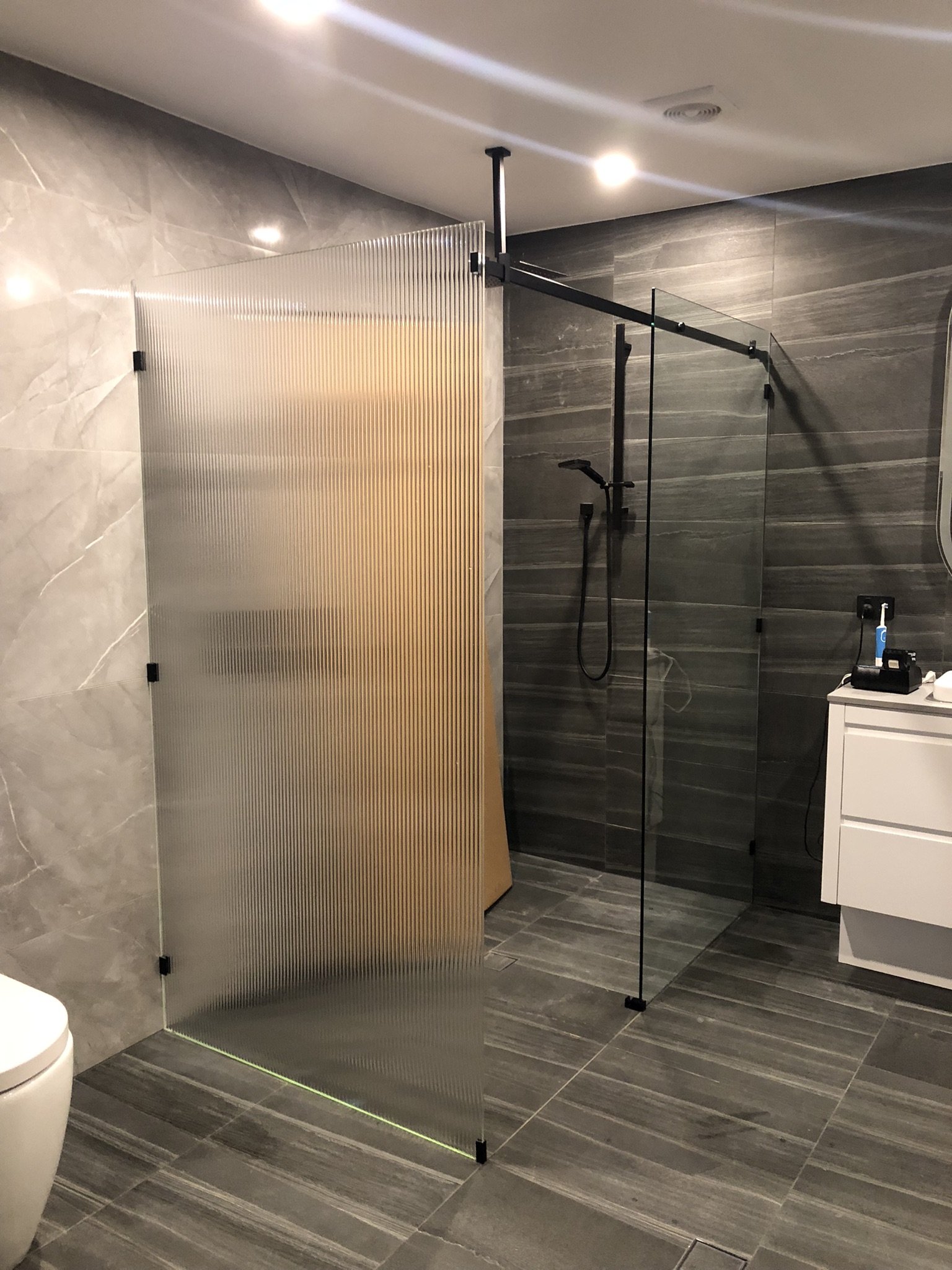 Modern bathroom with dark gray tiled walls and floor, a partial frosted glass shower enclosure with black hardware, and a white bathroom vanity with a toothbrush and electric shaver on top.